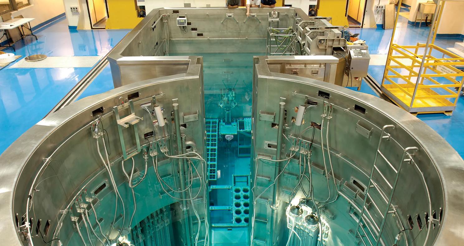 Interior of a nuclear research reactor facility showing a large stainless-steel reactor pool filled with clear blue water, with submerged reactor components, cables, and instrumentation visible, and technicians observing from a platform above.