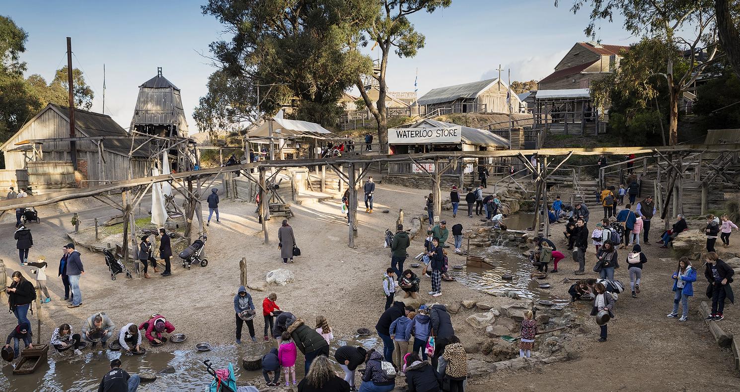 Students panning for gold at Sovereign Hill living museum in Ballarat, with historic buildings and mining structures in the background.