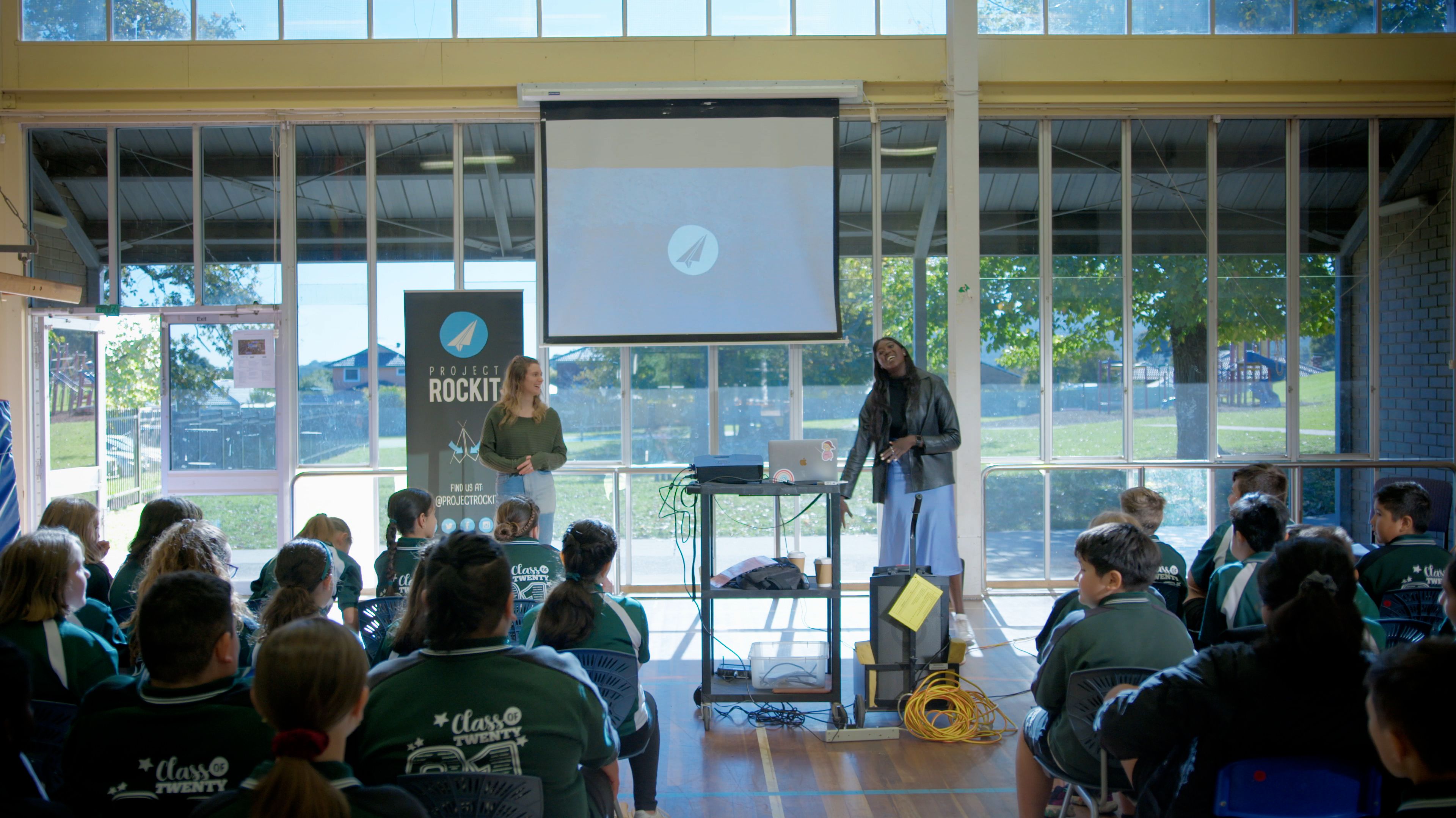 Two Project Rockit presenters run an interactive digital citizenship workshop for primary school students seated in a school hall, using a projector screen and presentation equipment while students listen and participate.