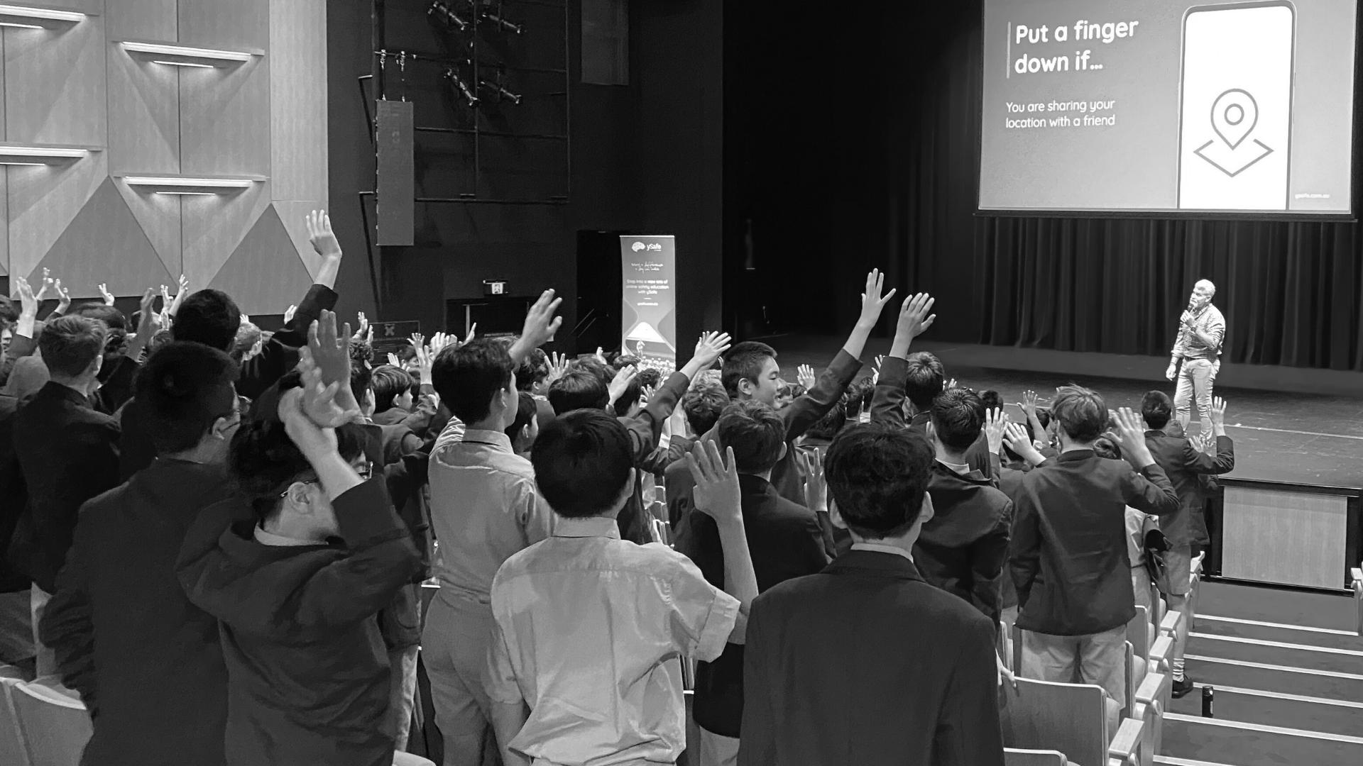Students in a school auditorium raise their hands during an online safety presentation on stage, with a slide reading “Put a finger down if… you are sharing your location with a friend.”
