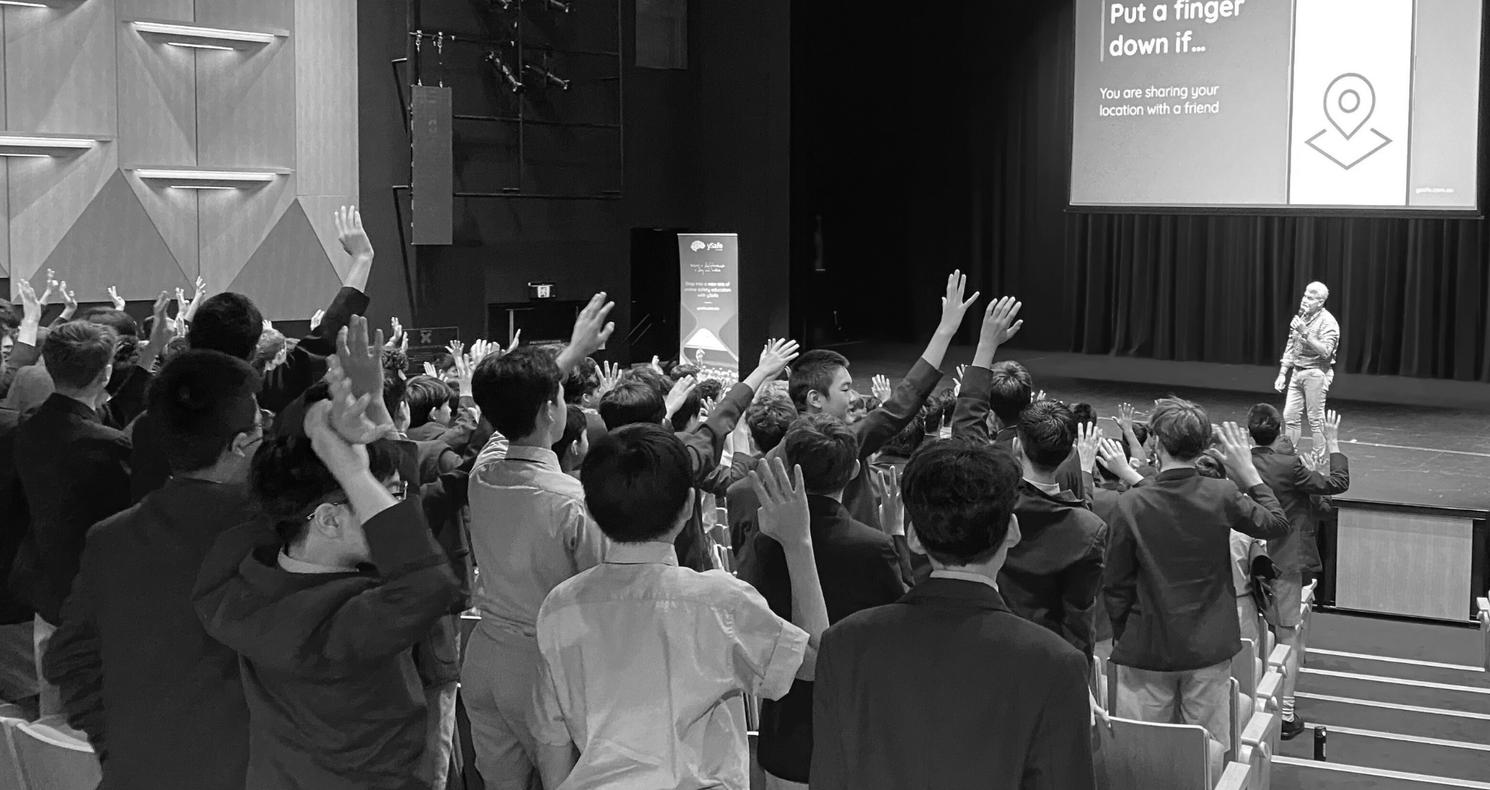 Students in a school auditorium raise their hands during an online safety presentation on stage, with a slide reading “Put a finger down if… you are sharing your location with a friend.”