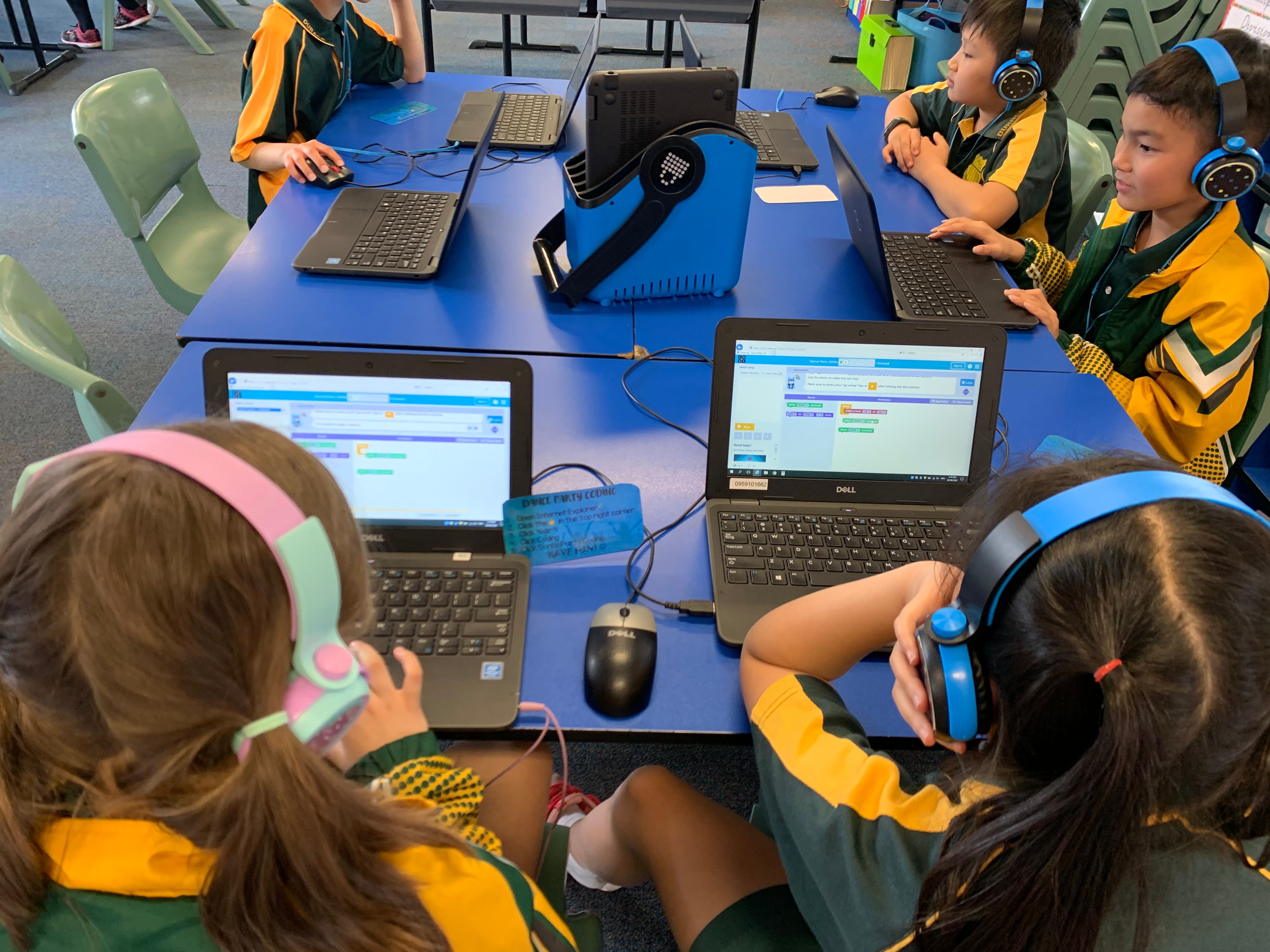 Primary school students in uniform wearing headphones and using laptops around a blue table in a classroom.