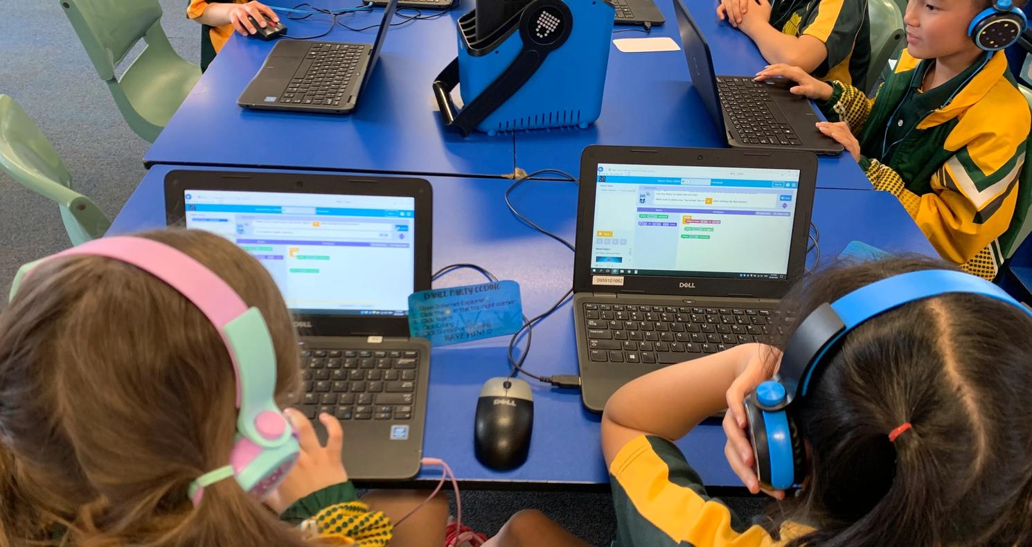 Primary school students in uniform wearing headphones and using laptops around a blue table in a classroom.