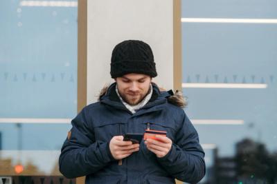 Homem com capuz e jaqueta segura o smartphone numa mão e o cartão de crédito na outra