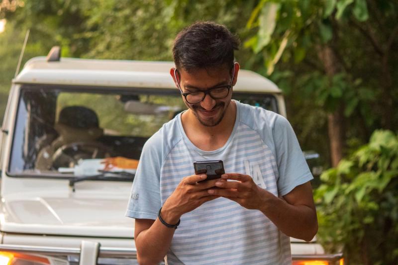 A man in a white crew neck t-shirt holding a black smartphone while on vacation.