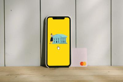 Smartphone mostra uma ilustração de banco digital ao lado de um cartão de crédito sobre uma mesa de madeira e parede branca ao fundo