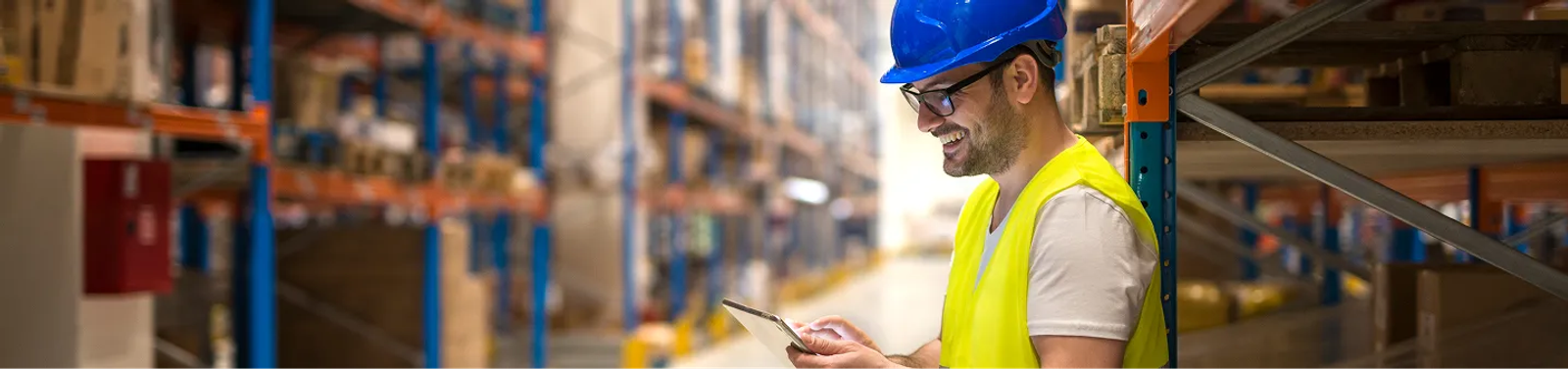 Operario de bodega con casco y chaleco reflectivo revisando información en una tablet entre estanterías industriales.