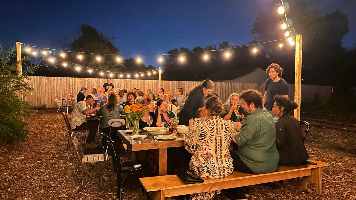 Guests share a long-table dinner in the garden at dusk, under strings of lights