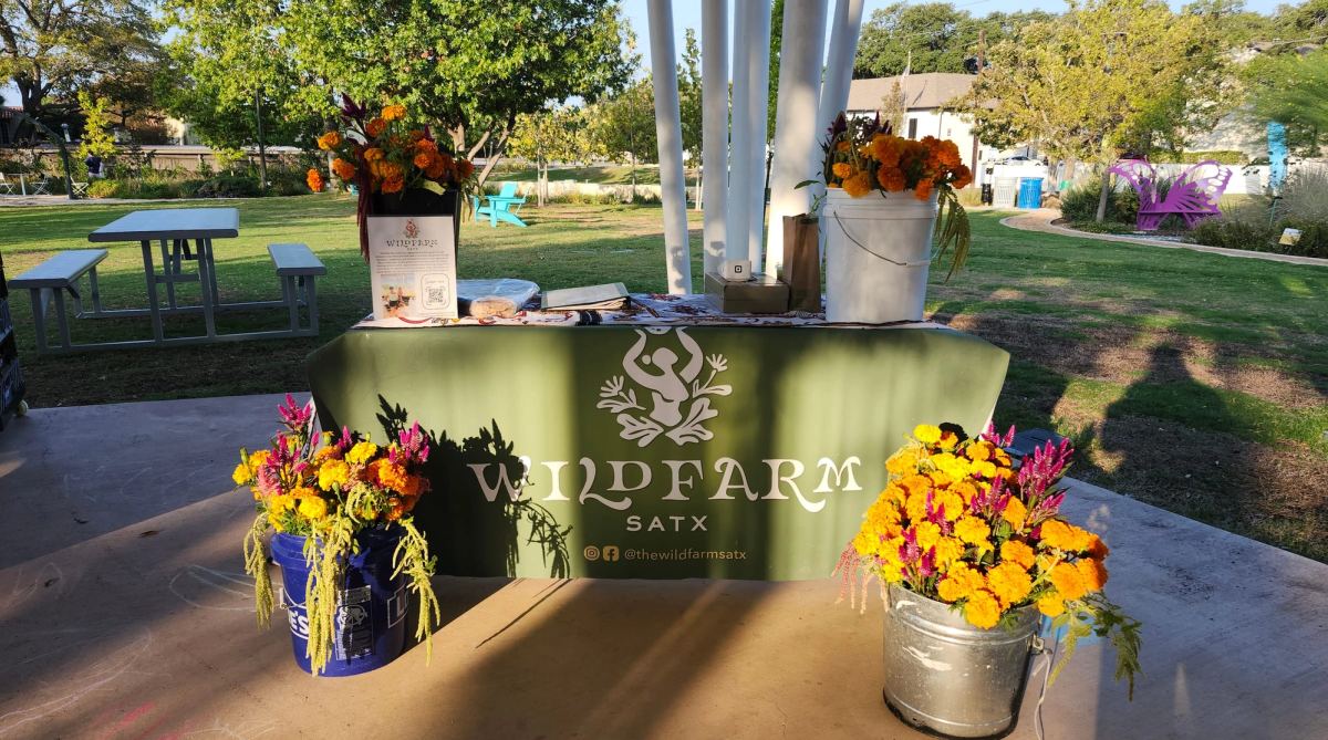 The Wild Farm’s market table under a park pavilion, draped in its green banner and set with buckets of marigolds