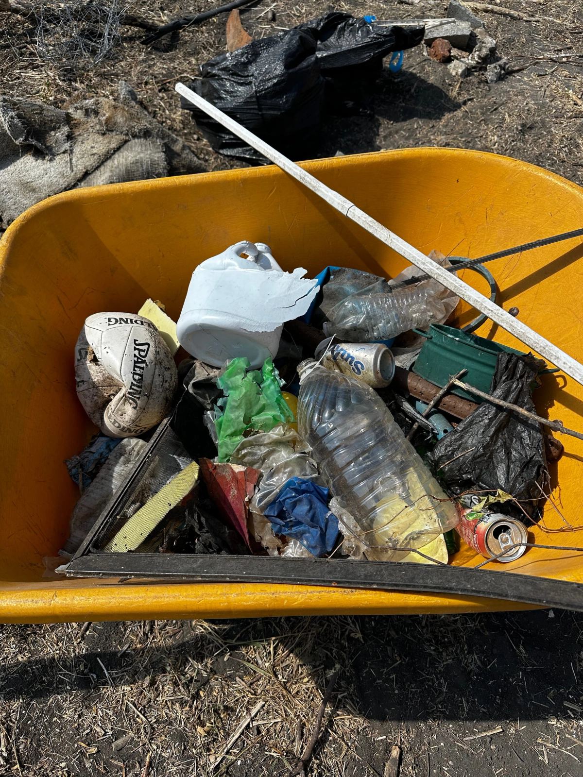 A yellow wheelbarrow full of trash cleared from the land: plastic bottles, cans, a deflated ball and scraps of plastic