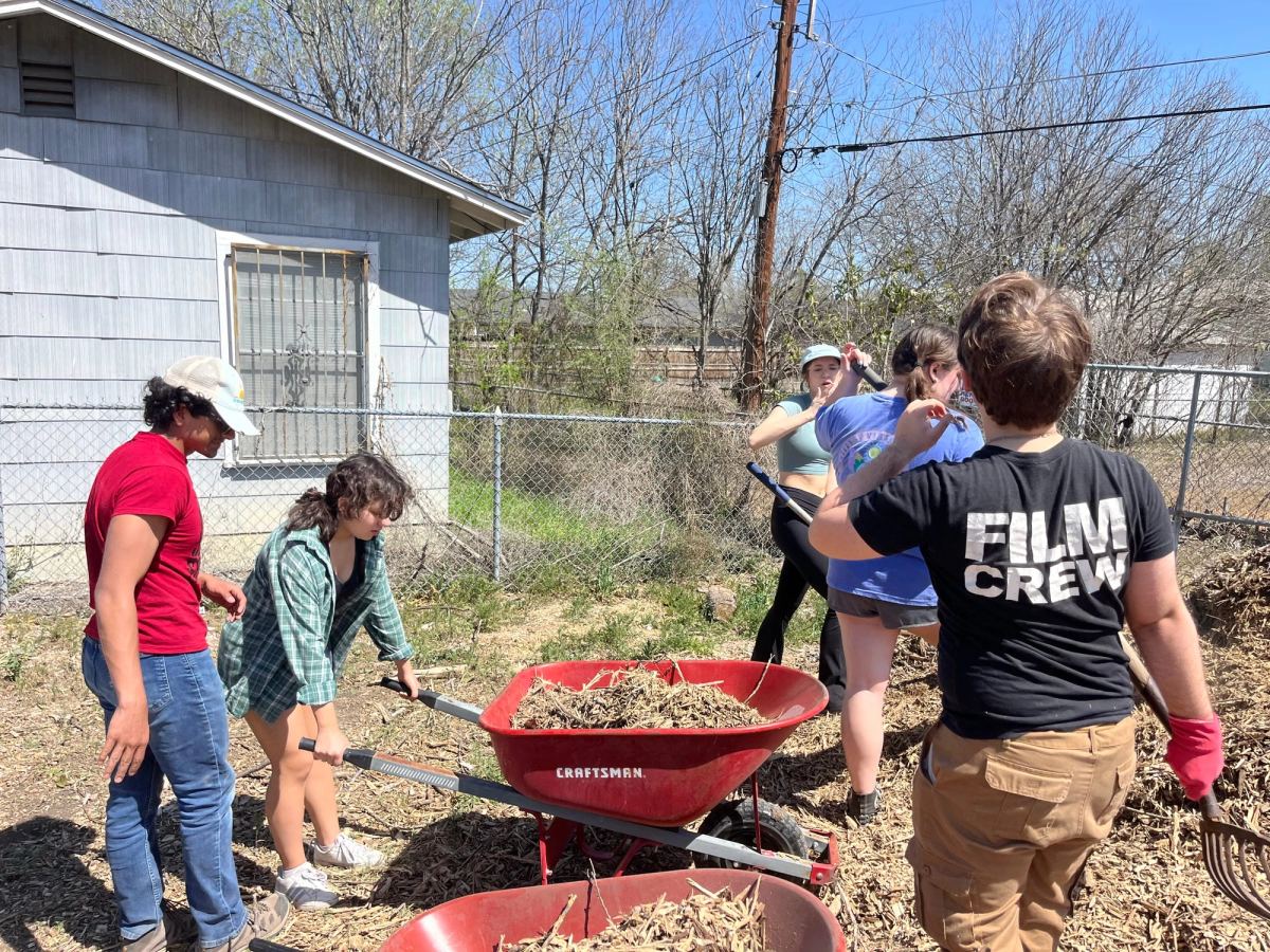 Volunteers fill red wheelbarrows with mulch beside a grey wooden building, one of them holding a pitchfork