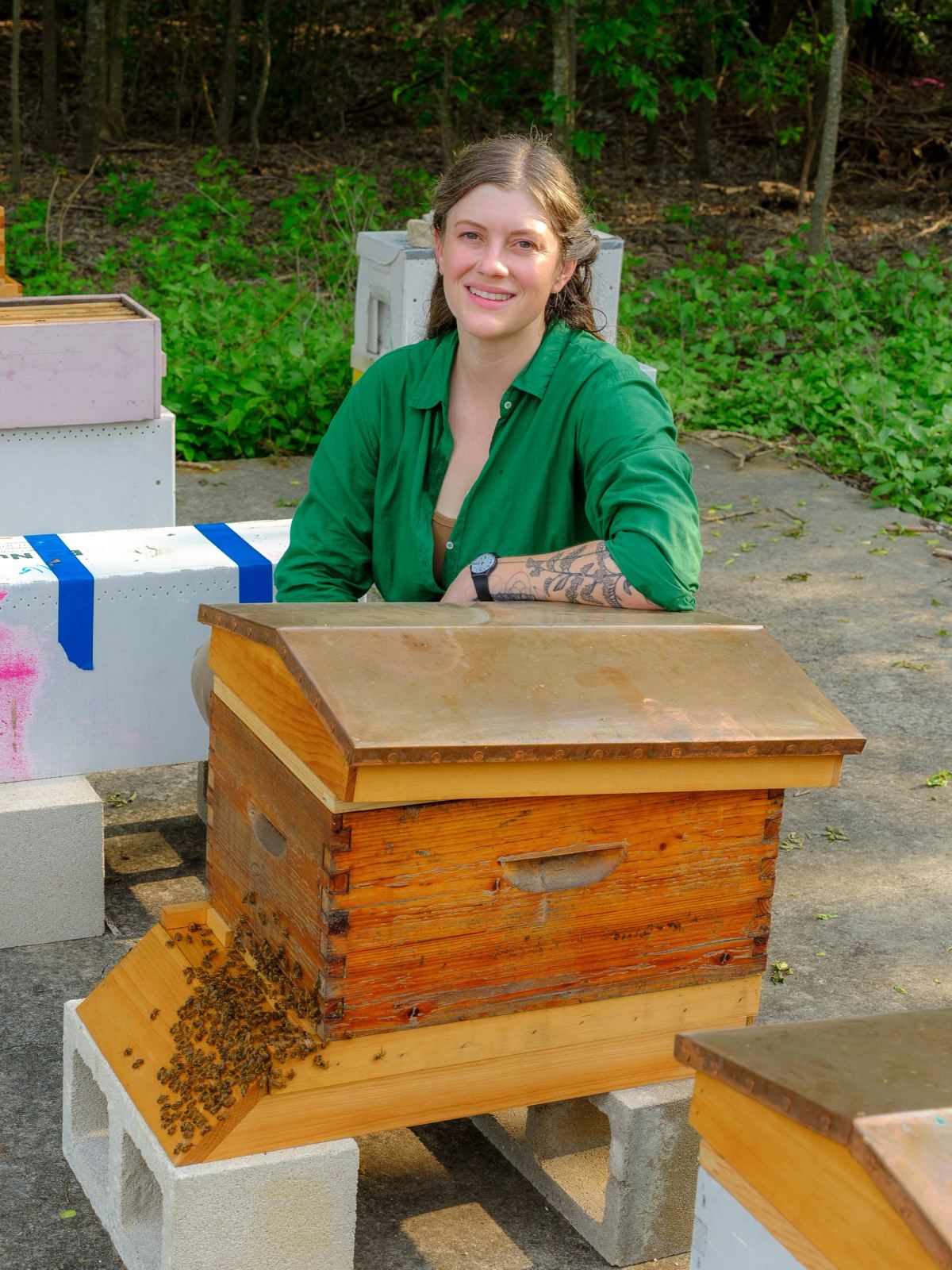 Cecile Parrish, head farmer and beekeeper, smiling behind a wooden beehive with bees gathered at its entrance