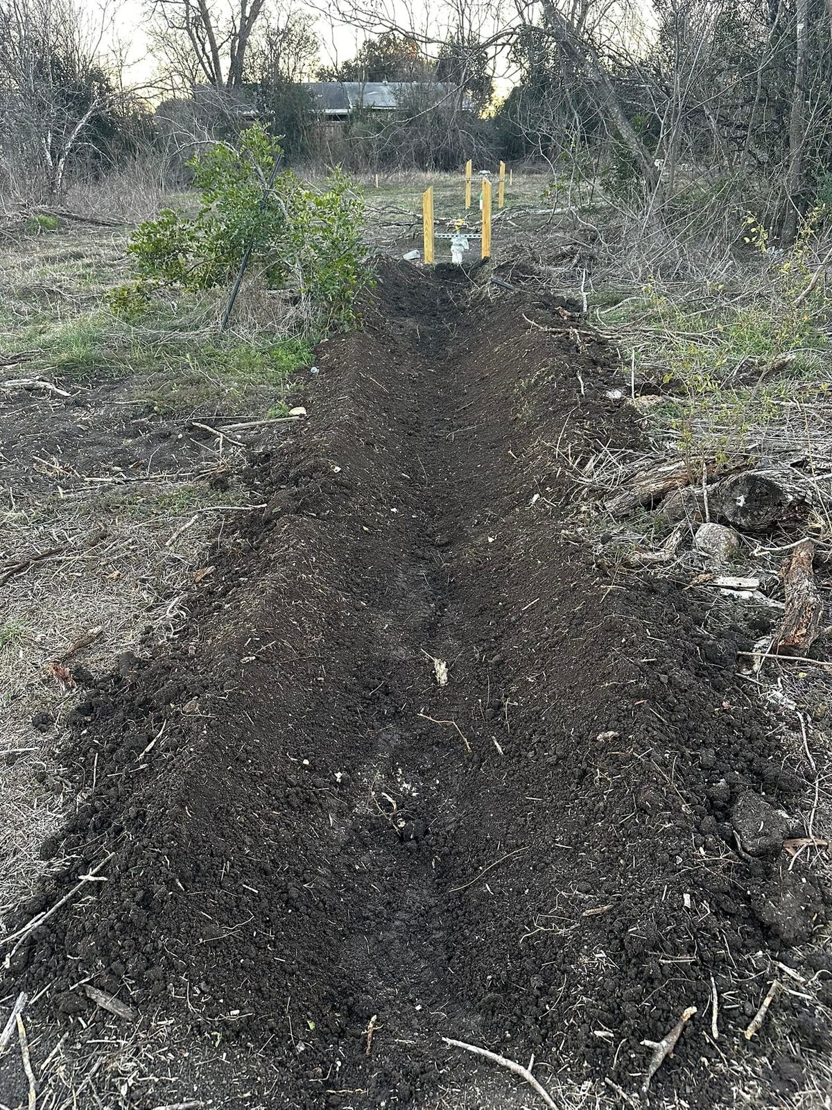 A long, freshly dug bed of dark soil running across the land toward two yellow posts
