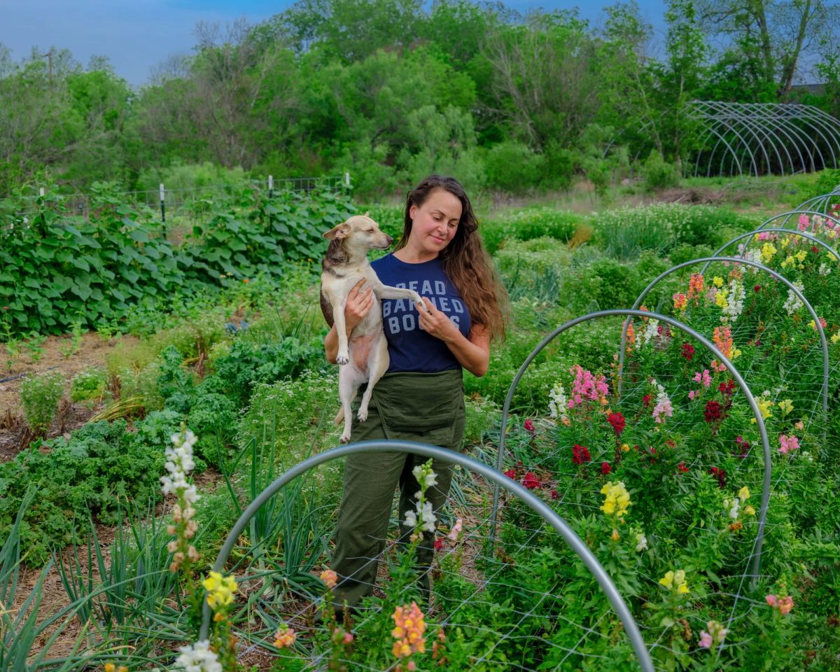 A woman stands among snapdragons in the garden, holding a small dog, with wire hoops arching over the rows
