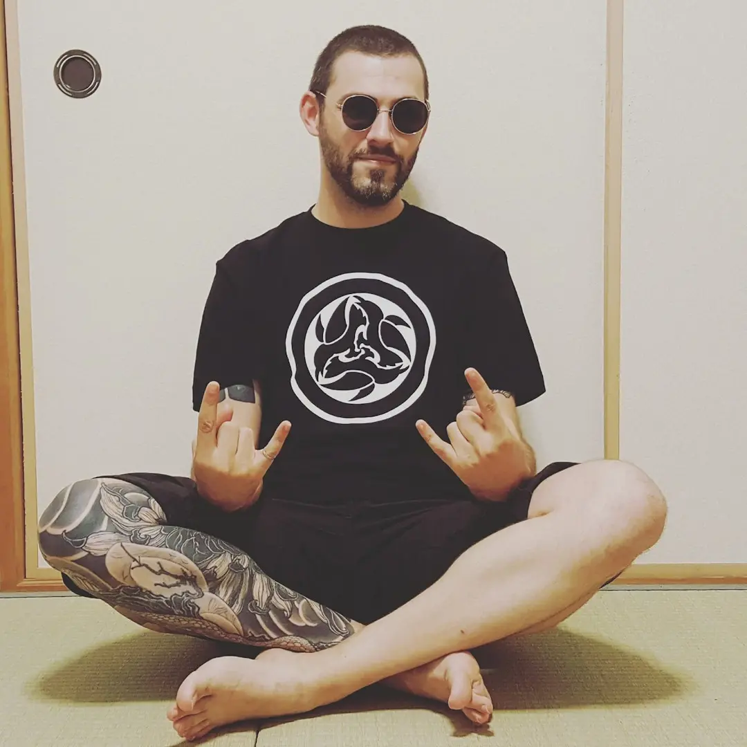 Ludovic Helme, also known as Shinobi, sits with crossed legs and hands in horns pose. He wears dark sunglasses.