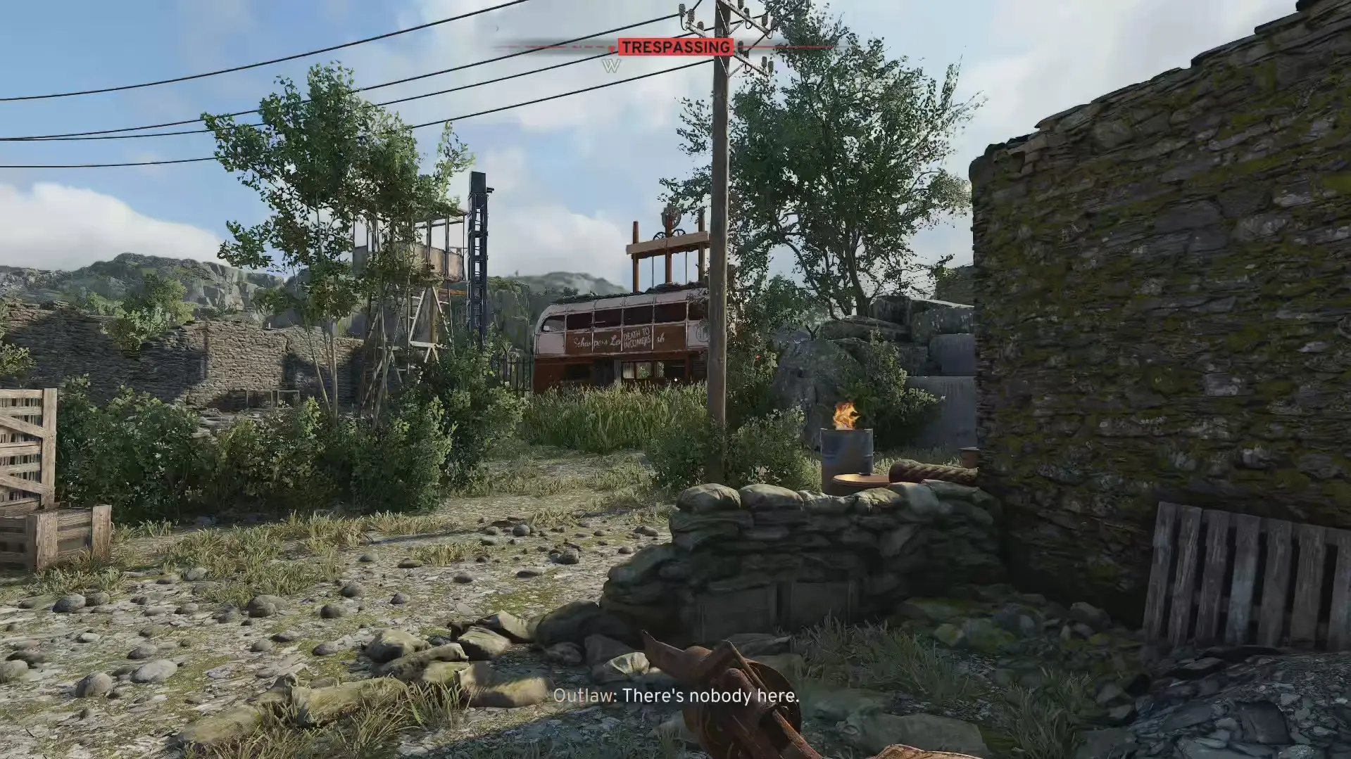 The player tresspasses in an outlaw camp with near a sandbag wall and small telephone pole. A double decker bus is visible beyond some foliage.