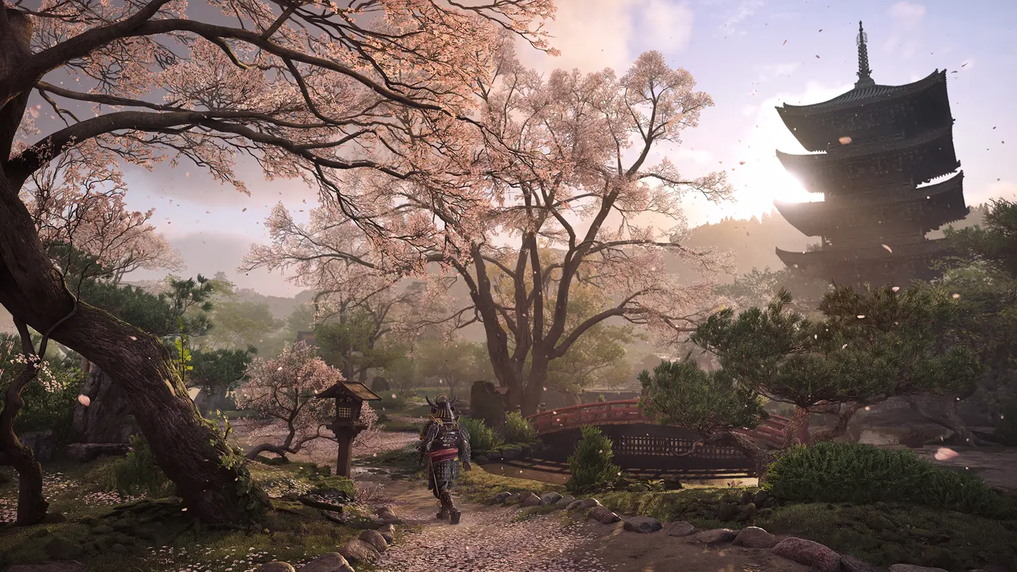 In the Assassin's Creed Shadows video game, Yasuke walks near a Japanese temple in a lush environment full of green and pink trees with pink petals covering the path