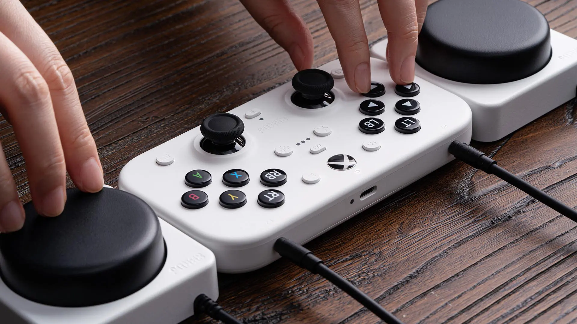 The 8BitDo Lite SE controller laying on a wooden surface. Two hands are interacting with the main controller and one of its Super Buttons