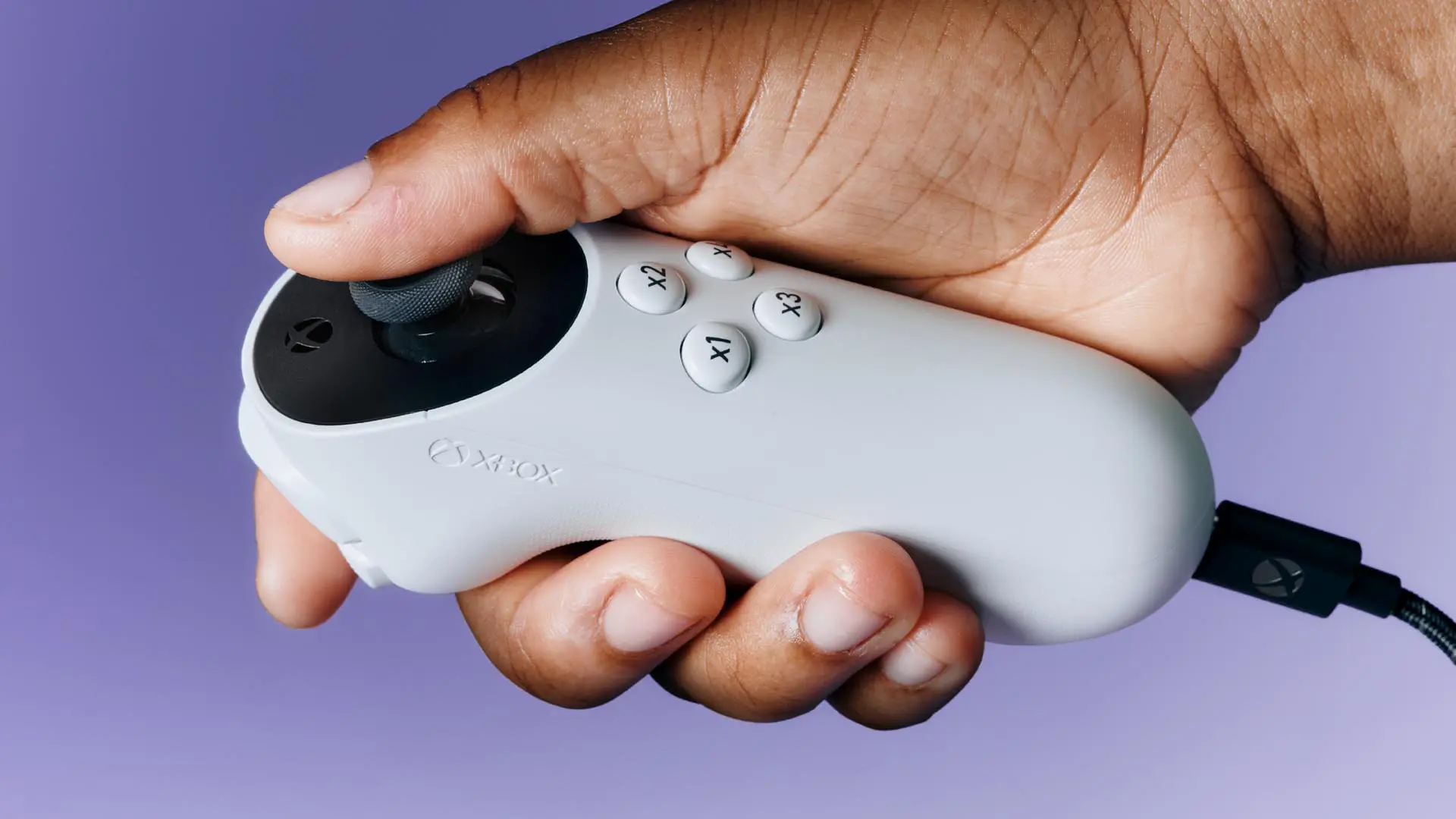 The Xbox Adaptive Joystick being held in a person's right hand against a light purple background