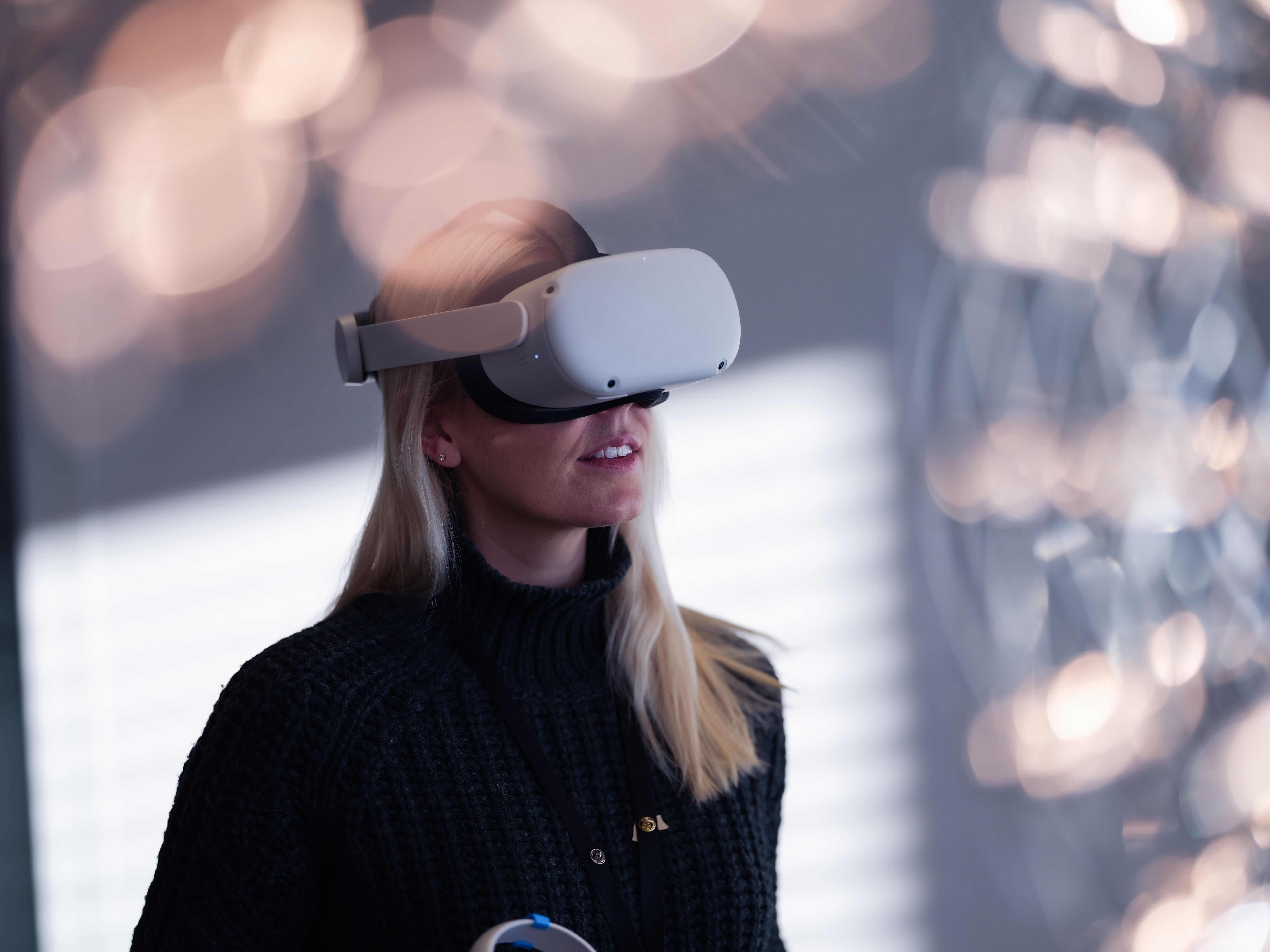 A woman with long blonde hair wearing a white VR headset, with blurred lights behind her.