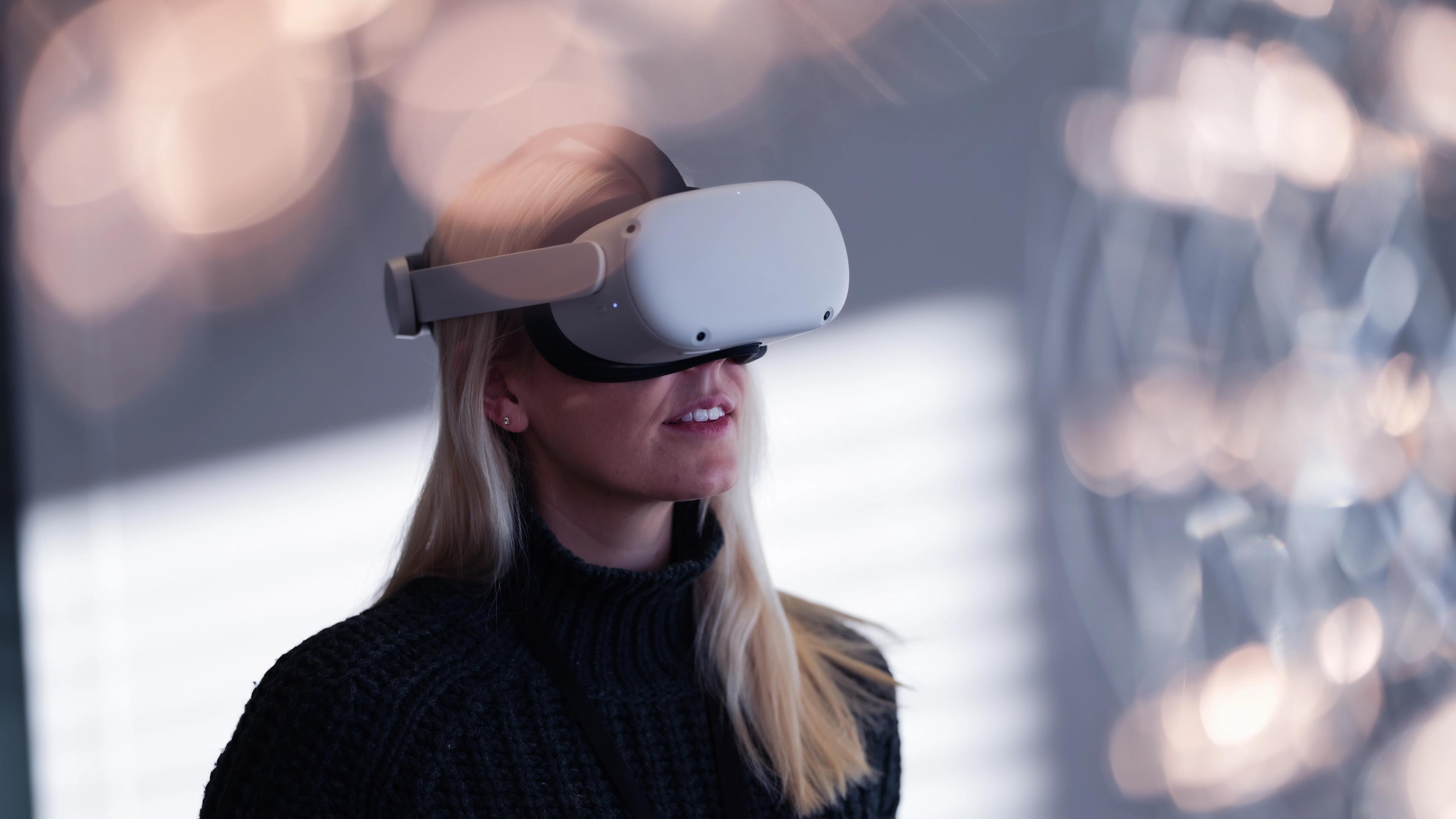 A woman with long blonde hair wearing a white VR headset, with blurred lights behind her.