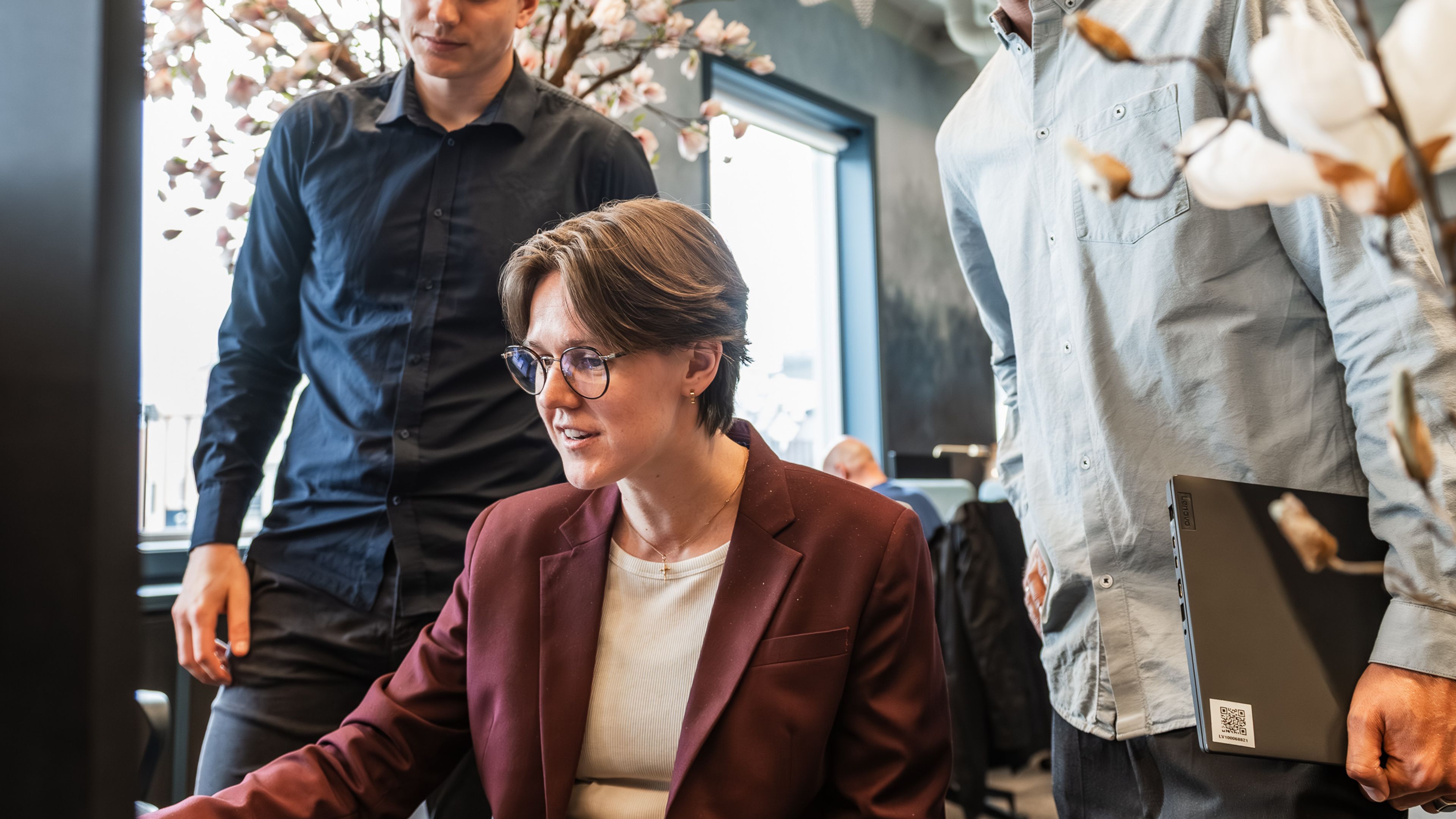 En kvinne i vinrød blazer sitter foran en skjerm. To menn står bak henne og følger med, i et kontorlandskap med synlige rør i taket.