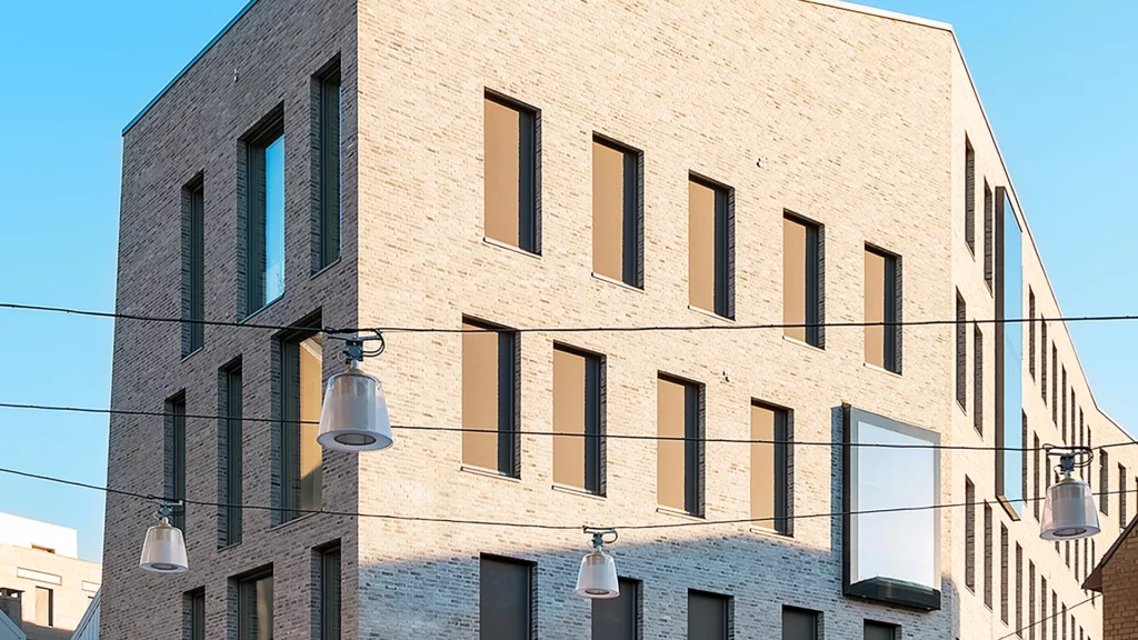 Hjørnet av Digs-bygget i lys tegl mot blå himmel. Gatelamper henger i vaiere foran fasaden.