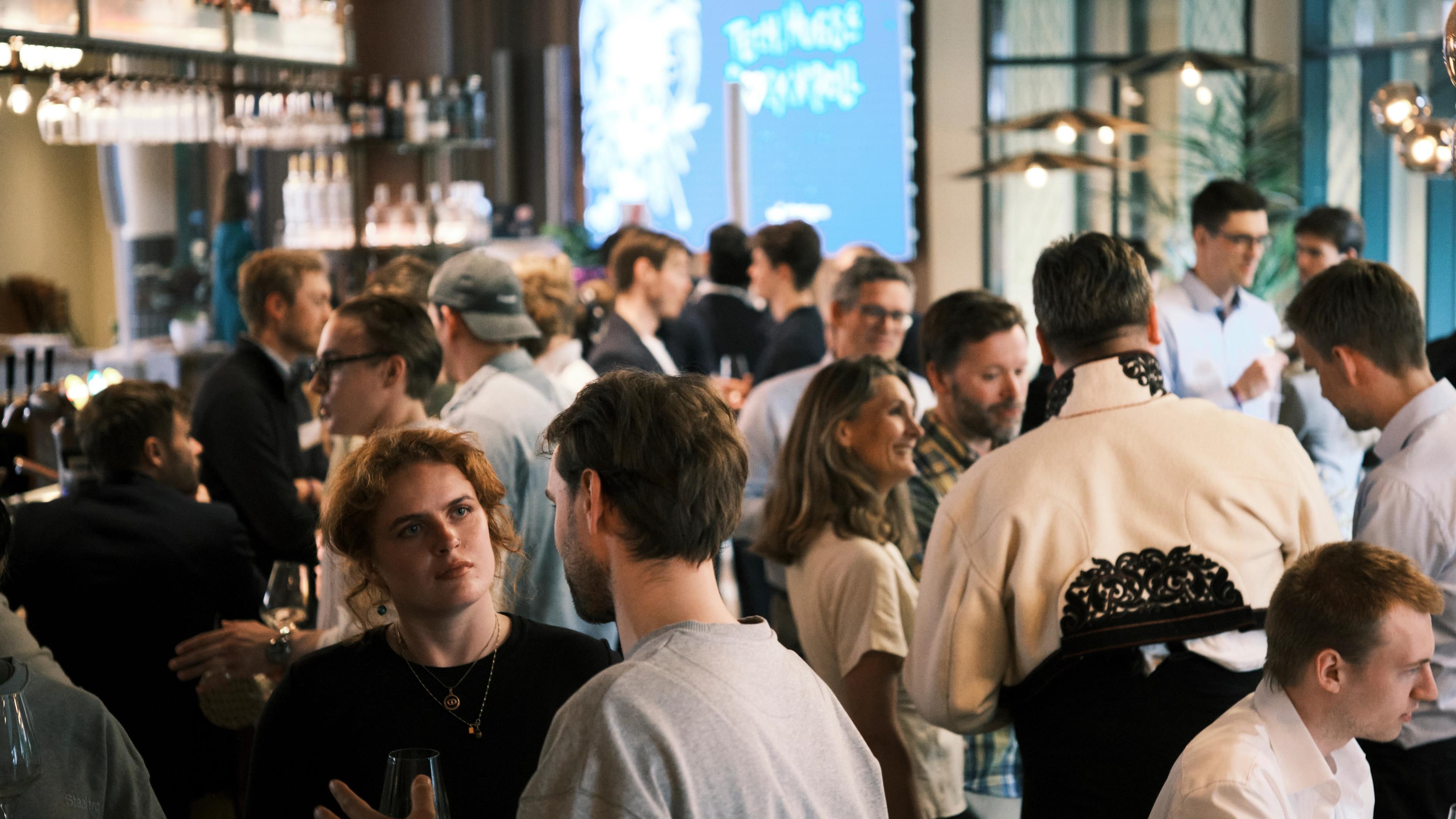 Tett i tett med folk som prater og holder glass i en bar. Én bærer en lys jakke med svarte broderier.