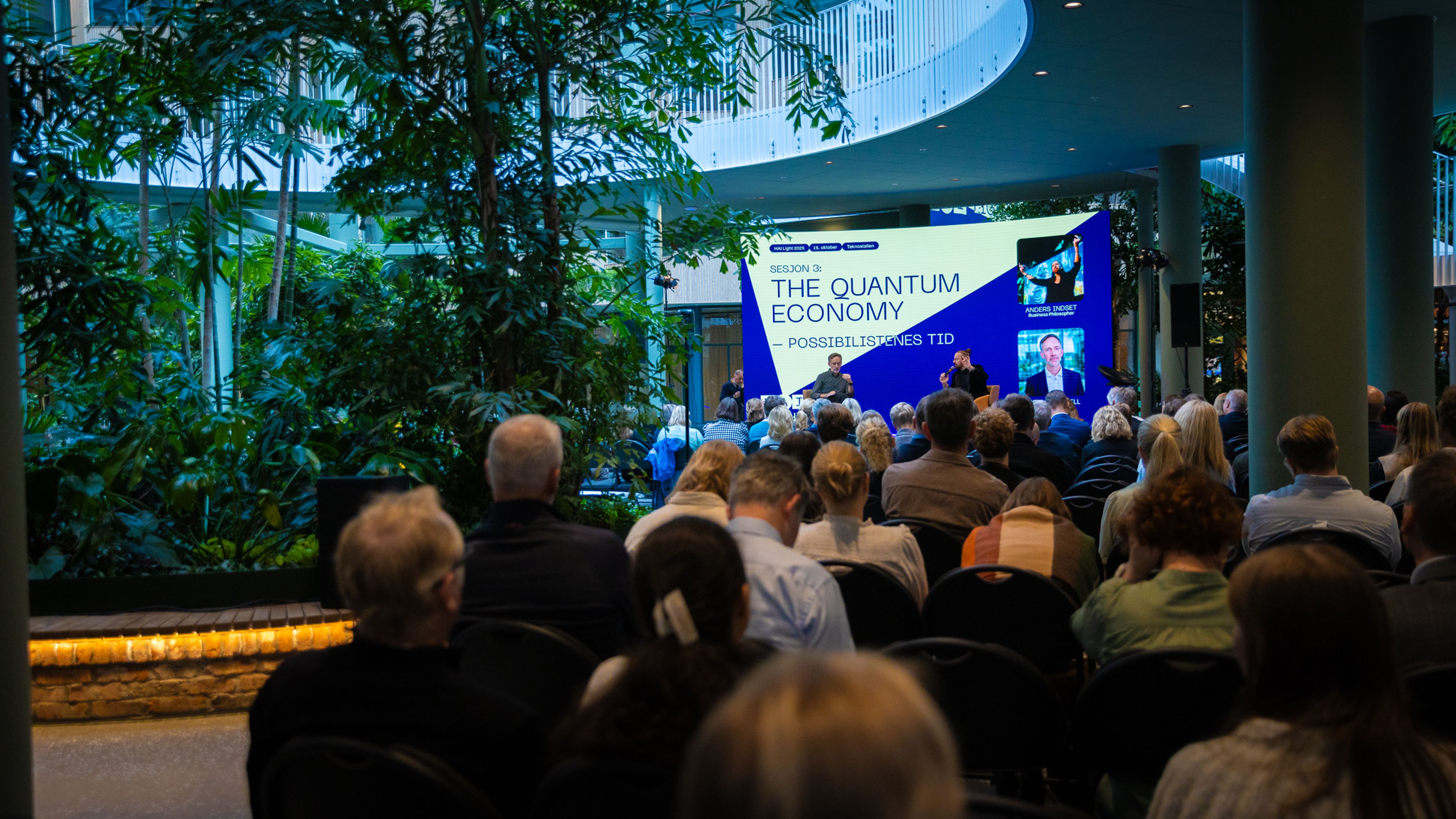 Publikum sett bakfra i en hall med store grønne planter. På scenen står en skjerm med teksten «The Quantum Economy – possibilismens tid».