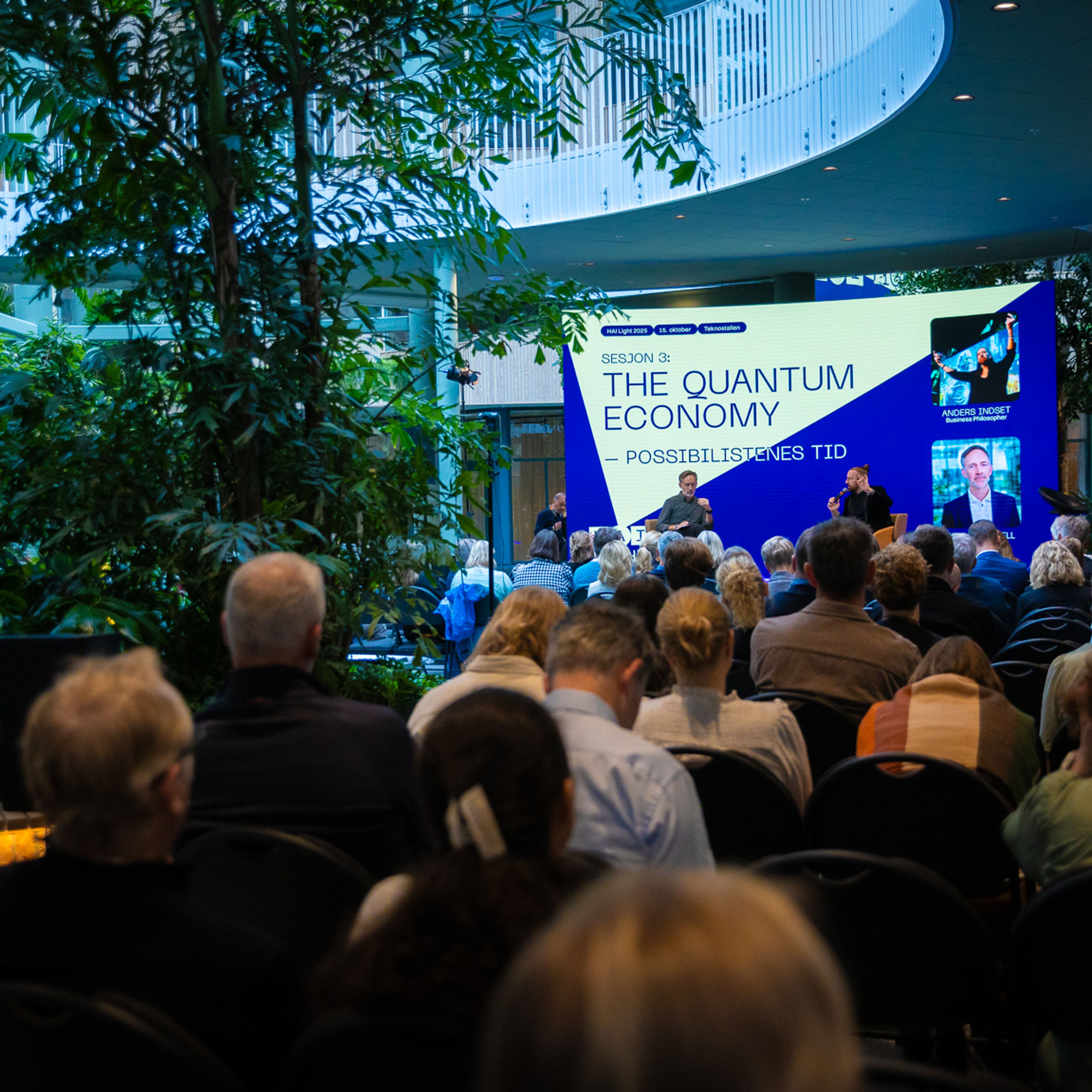 Publikum sett bakfra i en hall med store grønne planter. På scenen står en skjerm med teksten «The Quantum Economy – possibilismens tid».