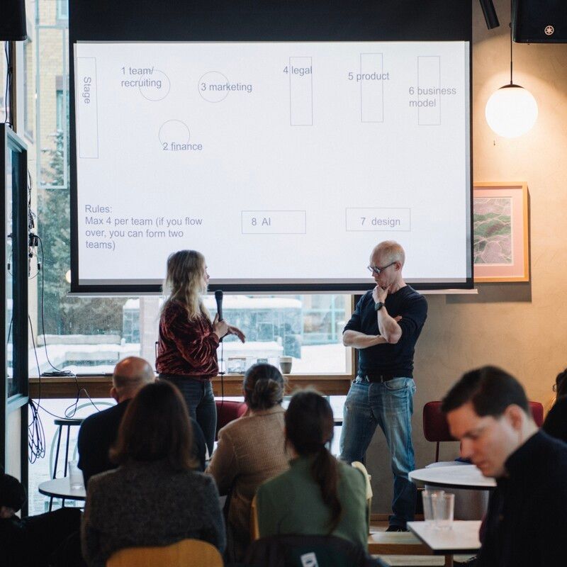 Two speakers, one holding a microphone, stand in front of a projected slide that lists team topics such as marketing, legal and AI. The audience sits at café tables.