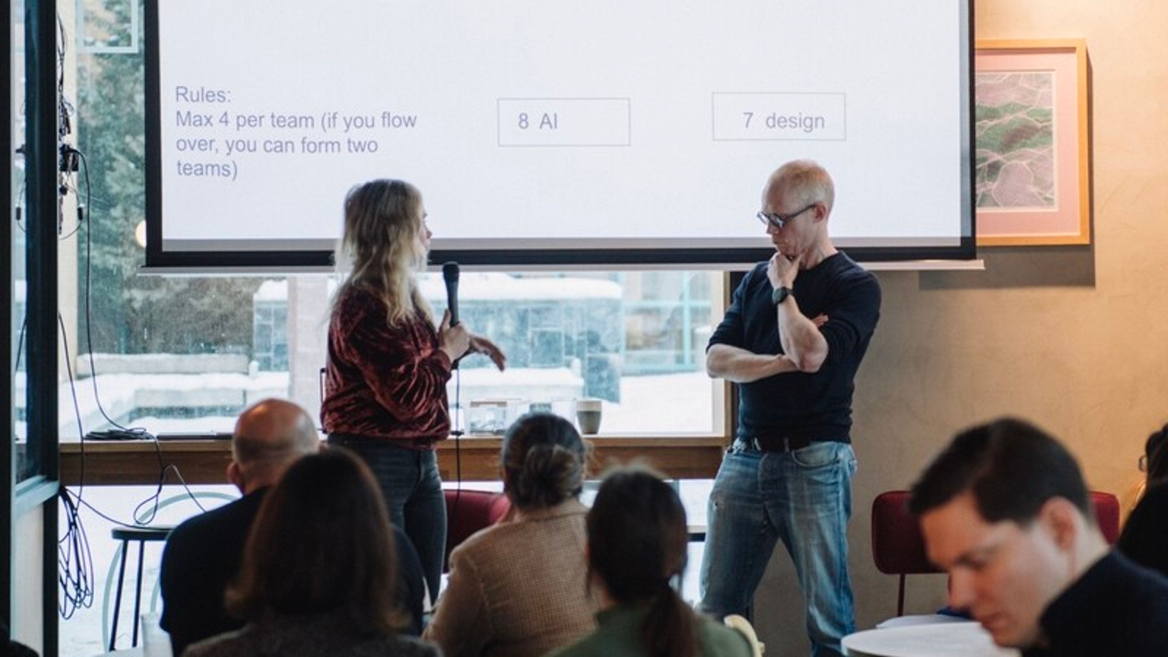 Two speakers, one holding a microphone, stand in front of a projected slide that lists team topics such as marketing, legal and AI. The audience sits at café tables.