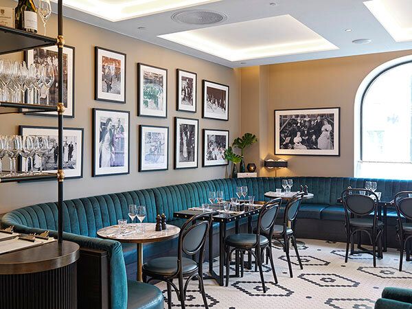 Restaurantlokale med buet, blågrønn fløyelssofa, små kafébord og svart-hvitt-fotografier på veggen.