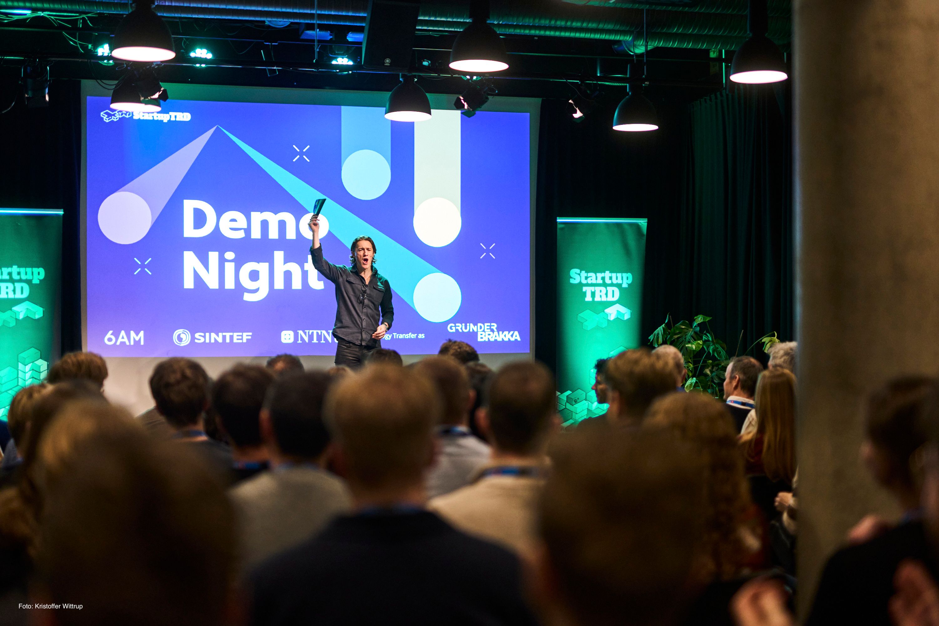 En programleder på scenen foran en skjerm med teksten «Demo Night», sett fra bakerst i en full sal.