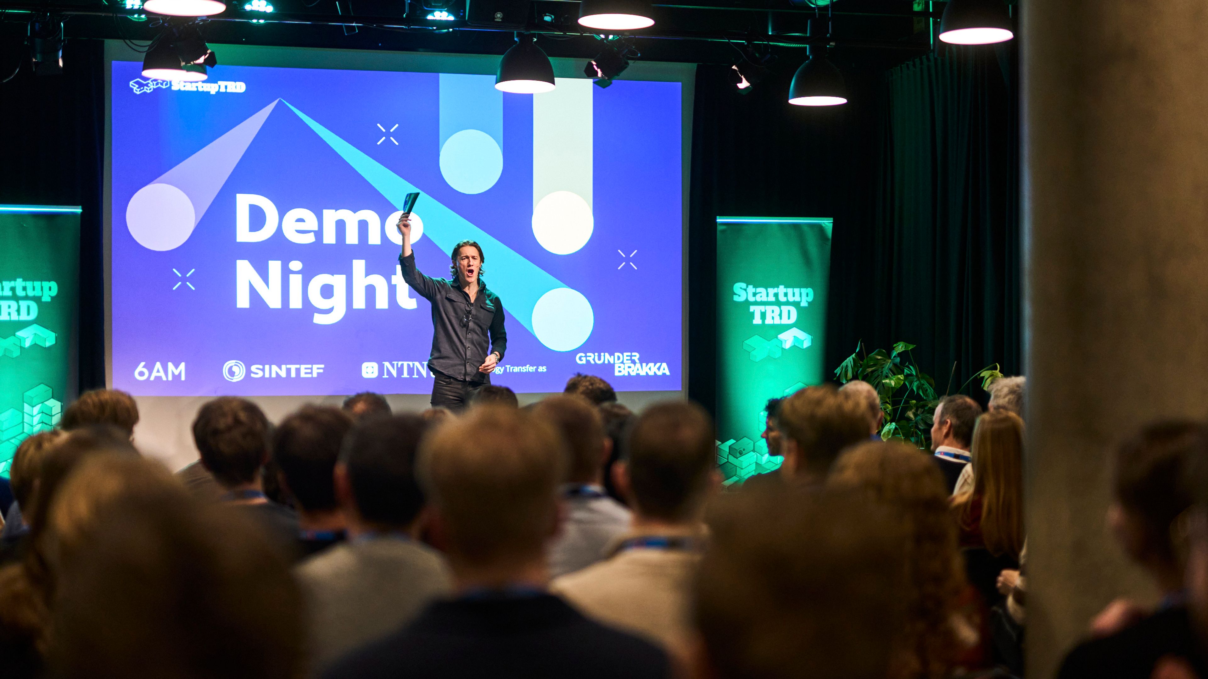 En programleder på scenen foran en skjerm med teksten «Demo Night», sett fra bakerst i en full sal.