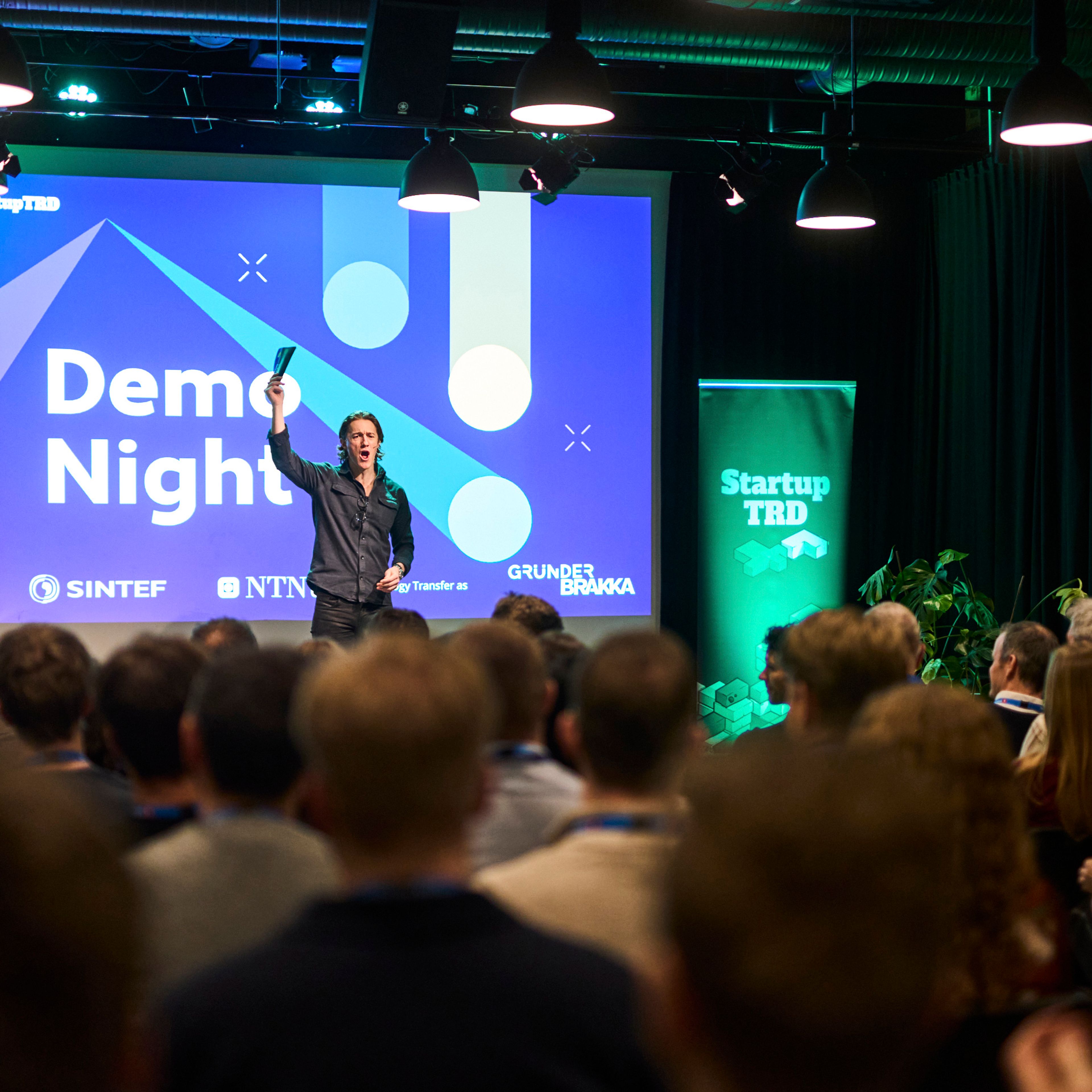 En programleder på scenen foran en skjerm med teksten «Demo Night», sett fra bakerst i en full sal.