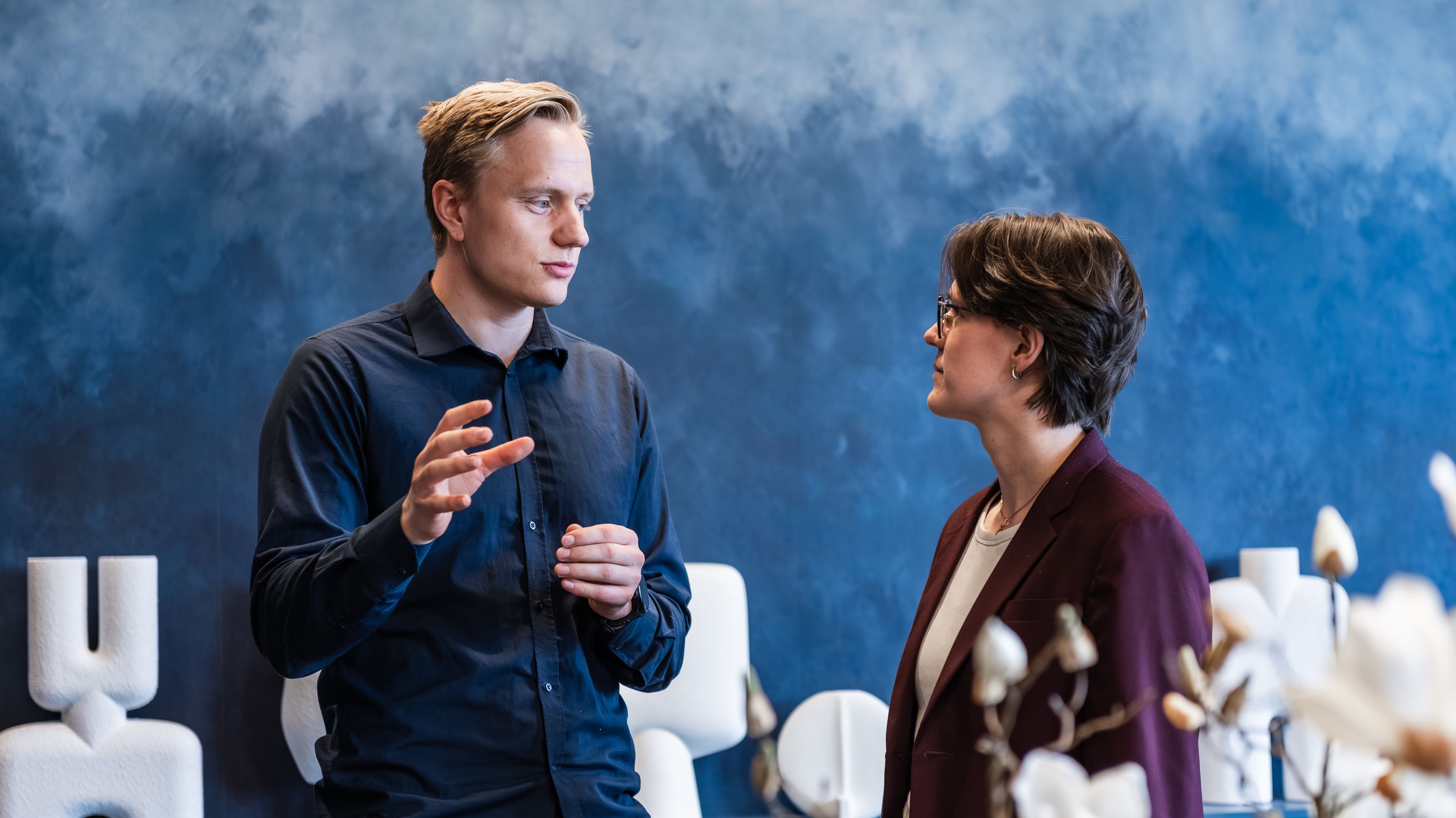 En mann i mørk skjorte forklarer noe med hendene til en kvinne i vinrød blazer, foran en blåmalt vegg.