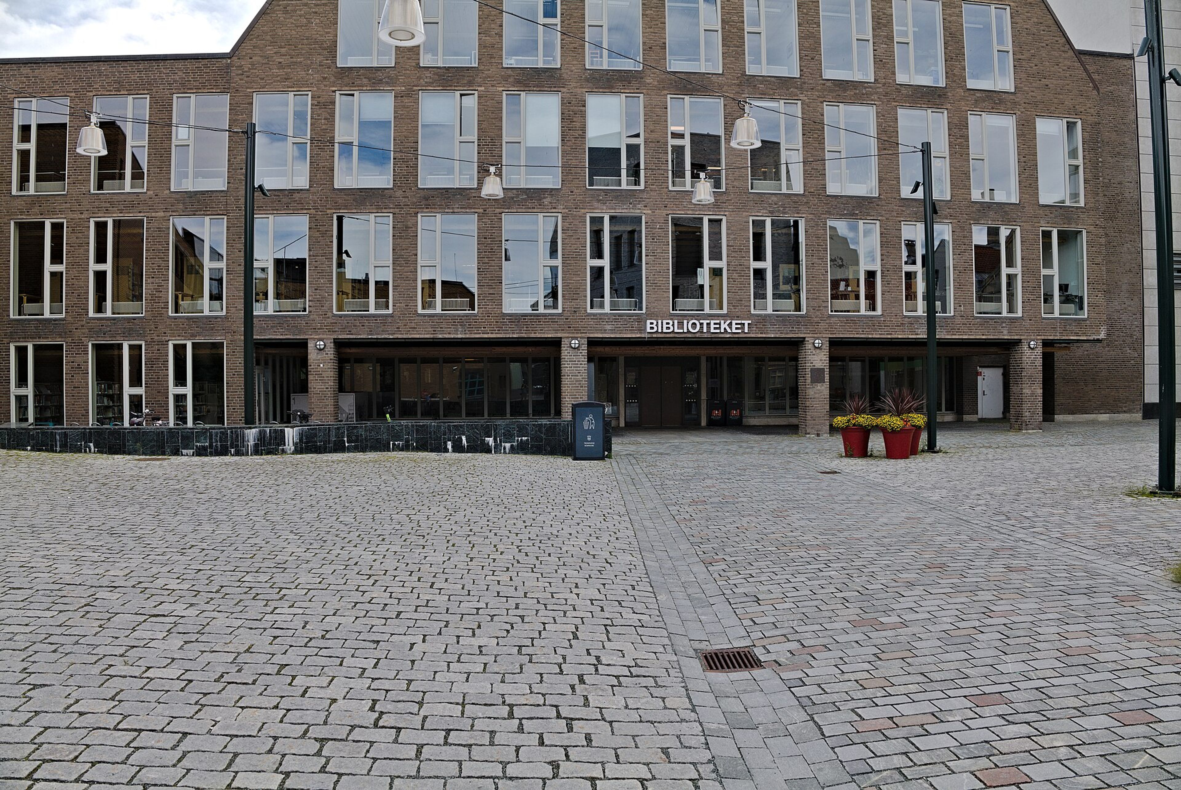 Den brosteinsbelagte plassen foran en teglbygning med skiltet «Biblioteket» over inngangen.