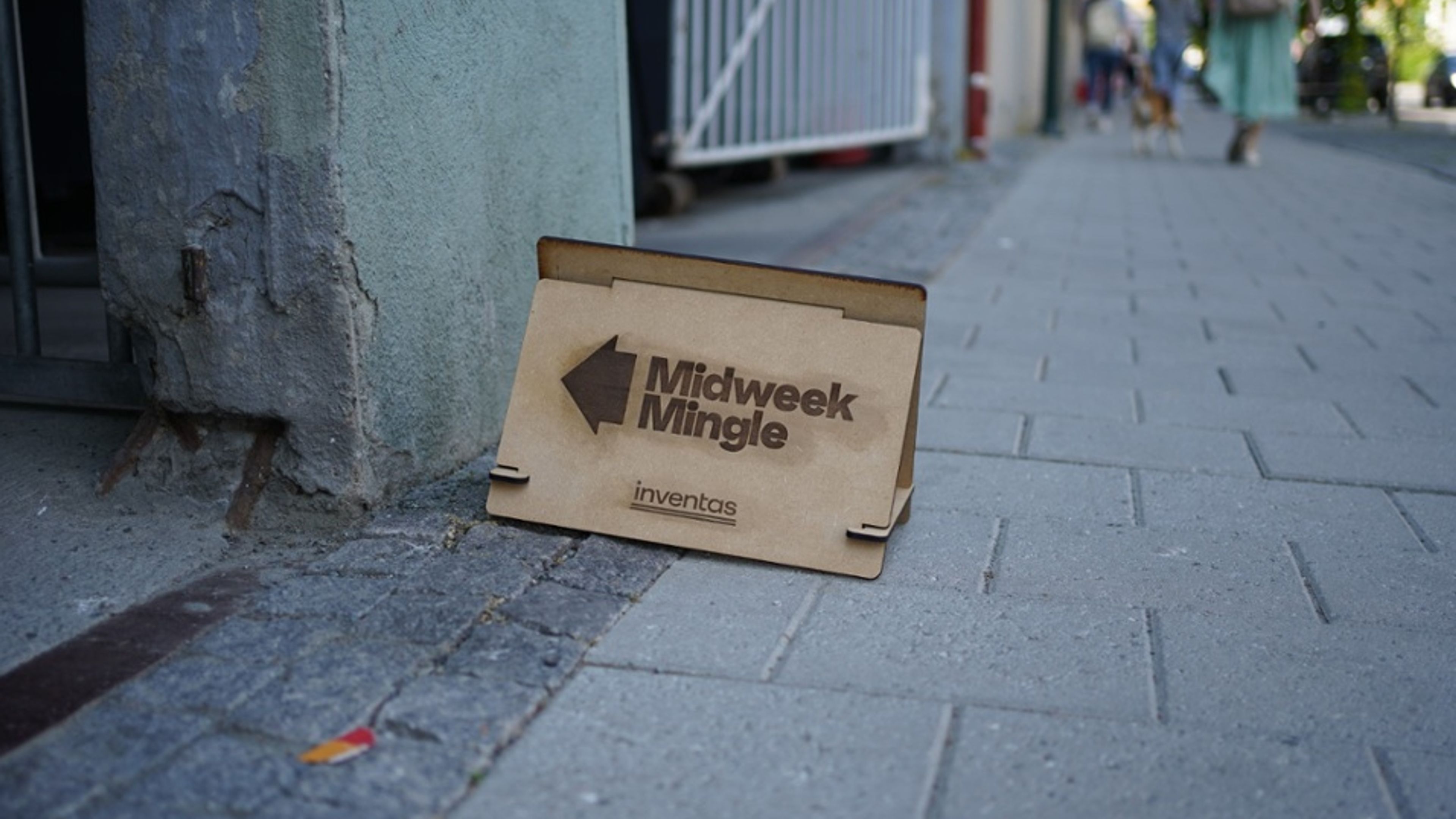 A wooden Midweek Mingle sign on a pavement pointing to the entrance