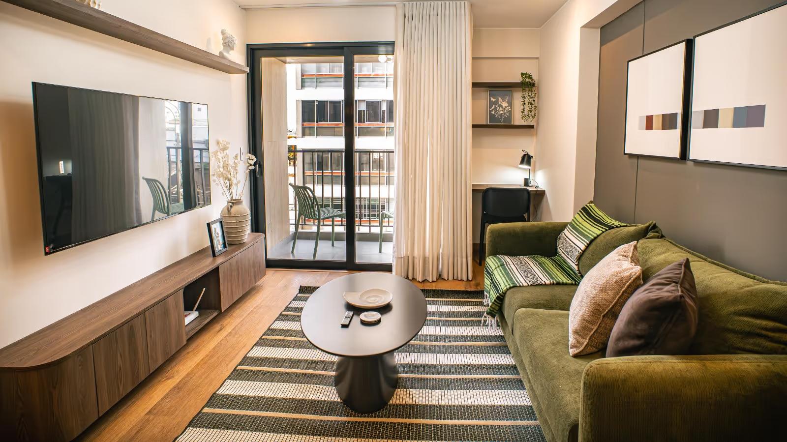 Compact apartment living room with olive sofa, striped rug, balcony doors and wall-mounted TV