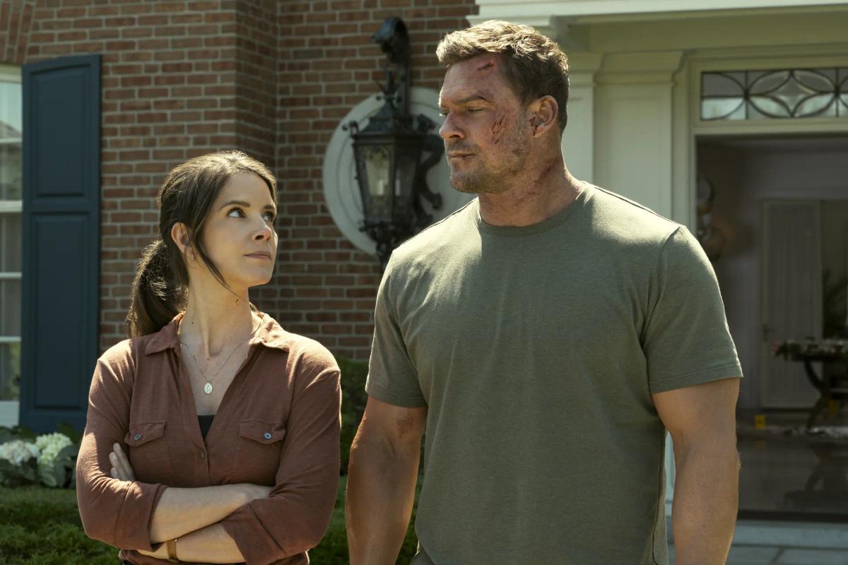 Sonya Cassidy as Susan Duffy stands with her arms folded, looking up at Alan Ritchson as Jack Reacher, whose face is cut and bruised, outside a brick house with an evidence marker on the floor behind them.