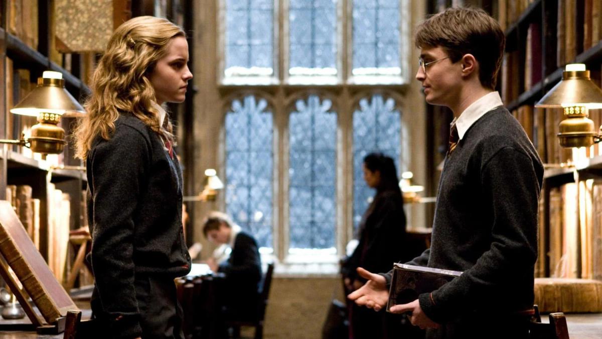 Emma Watson as Hermione Granger facing Daniel Radcliffe as Harry Potter, who is holding books in the Hogwarts library