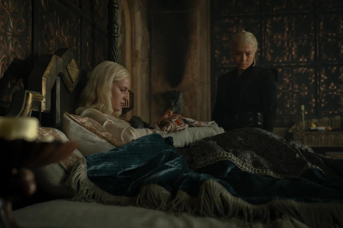 Phia Saban as Helaena Targaryen sews in bed while Emma D’Arcy as Rhaenyra Targaryen stands over her in black