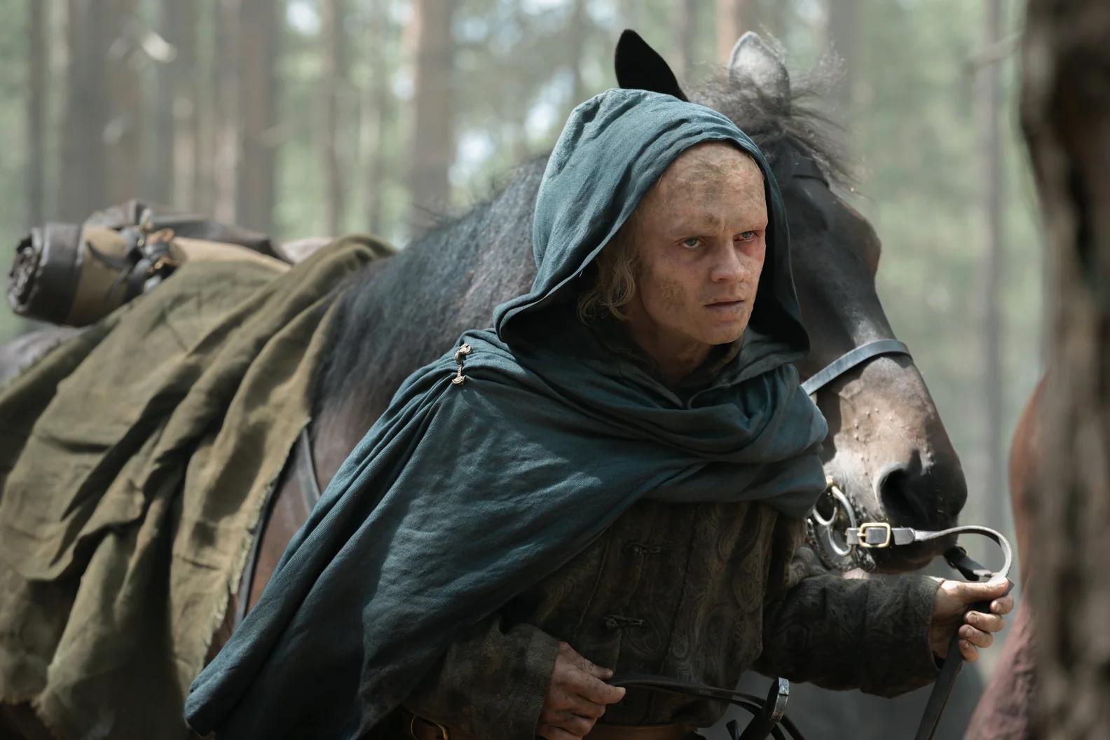 Tom Glynn-Carney as Aegon II Targaryen, hooded and scarred, leading a horse through woodland