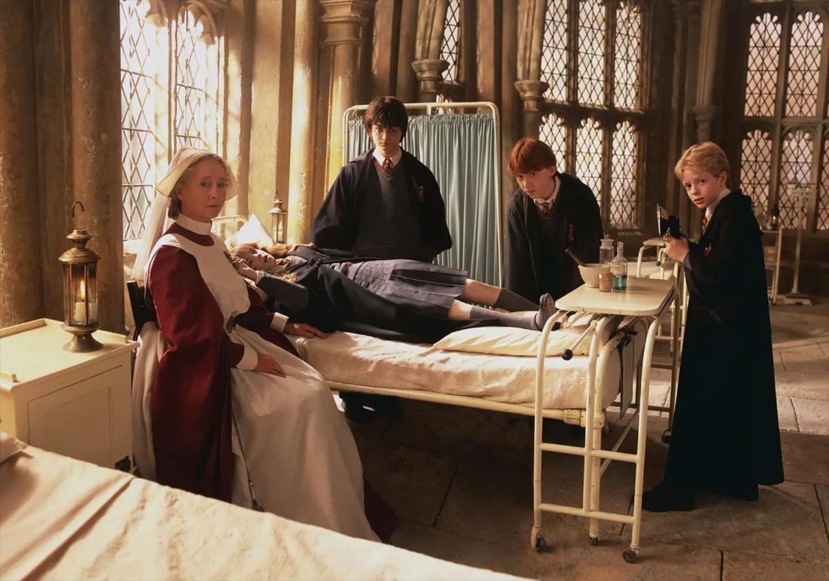 Emma Watson as Hermione Granger lies petrified in a hospital bed while Daniel Radcliffe as Harry Potter and Rupert Grint as Ron Weasley stand beside her.