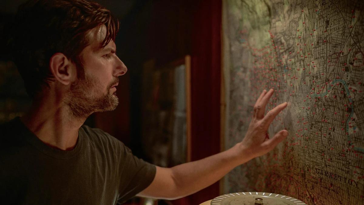 Adam Scott as Tom Kennedy reaching towards a wall map of Garretton covered in red pins in The Whisper Man.