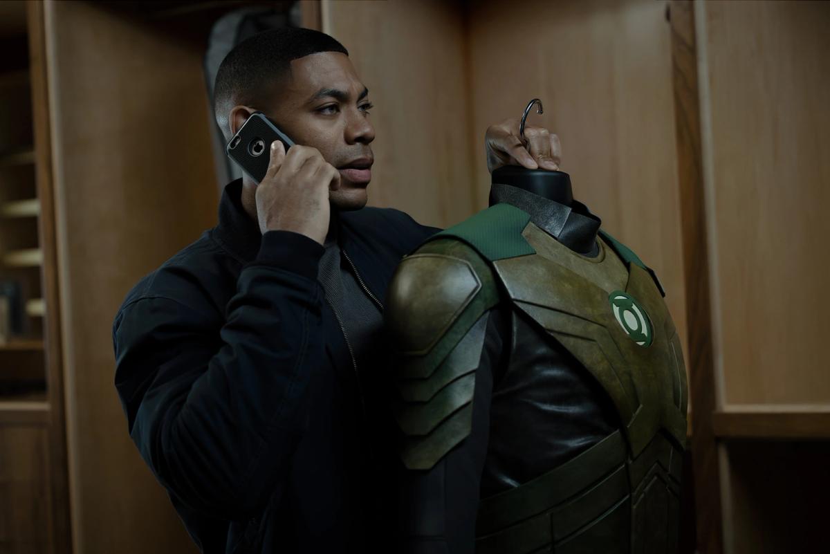 Aaron Pierre as John Stewart on the phone, holding a Green Lantern uniform on a hanger in a locker room.