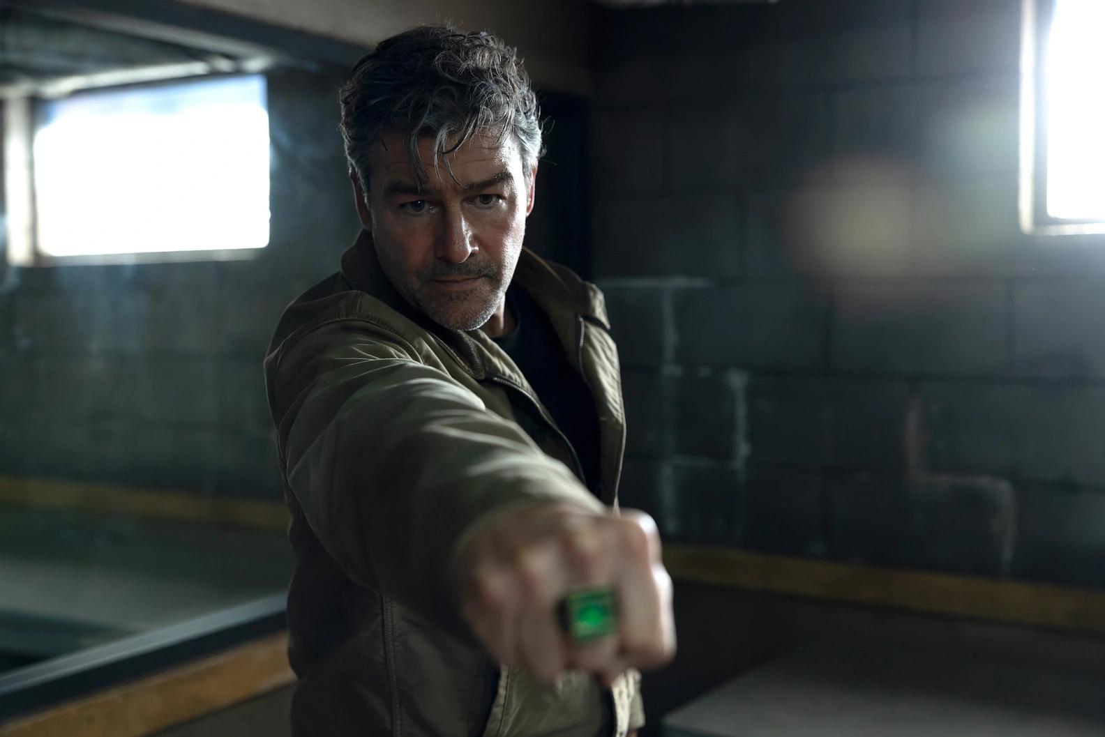 Kyle Chandler as Hal Jordan in a bare concrete room, arm extended towards the camera with a glowing green power ring on his hand.