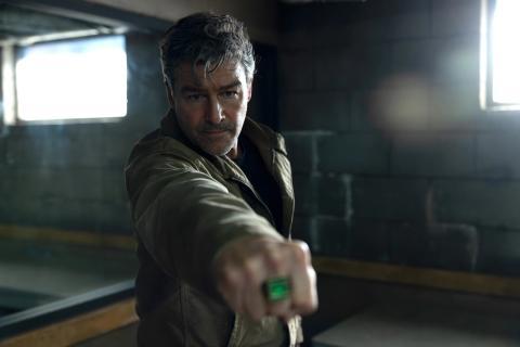 Kyle Chandler as Hal Jordan in a bare concrete room, arm extended towards the camera with a glowing green power ring on his hand.