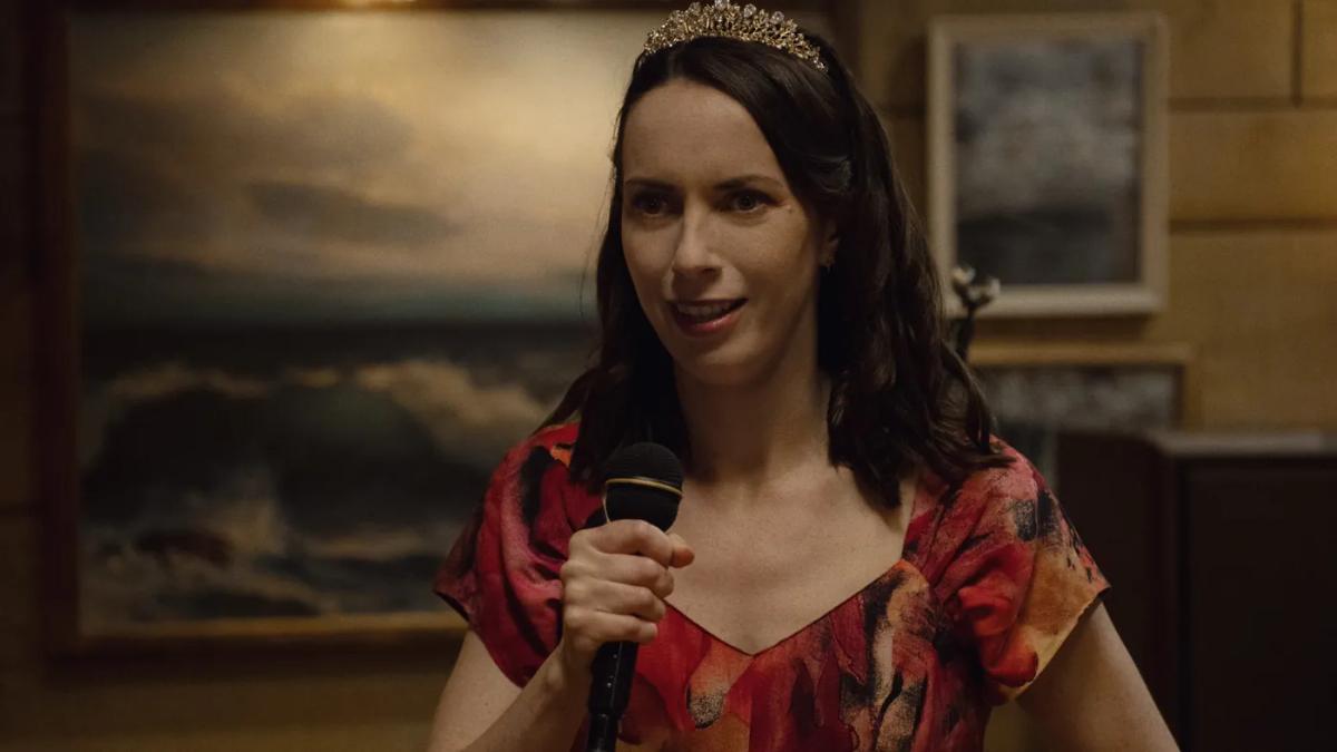 Kate O'Flynn as Patricia holds a microphone and addresses a room, wearing a gold tiara and a red and orange floral dress, a seascape painting on the wall behind her.